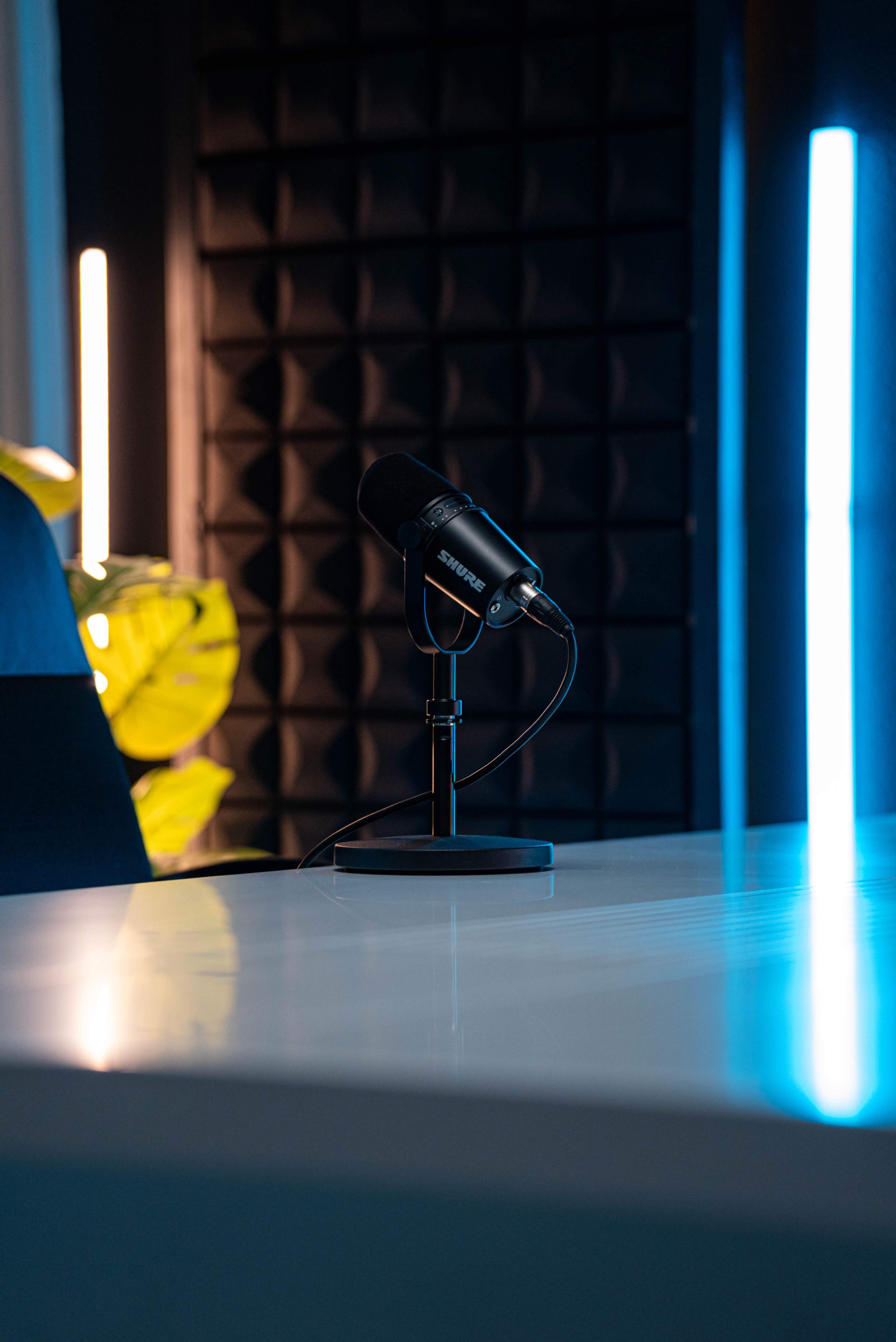 A black microphone stands on a sleek desk, with a textured black wall and dimly lit blue and white light fixtures in the background.