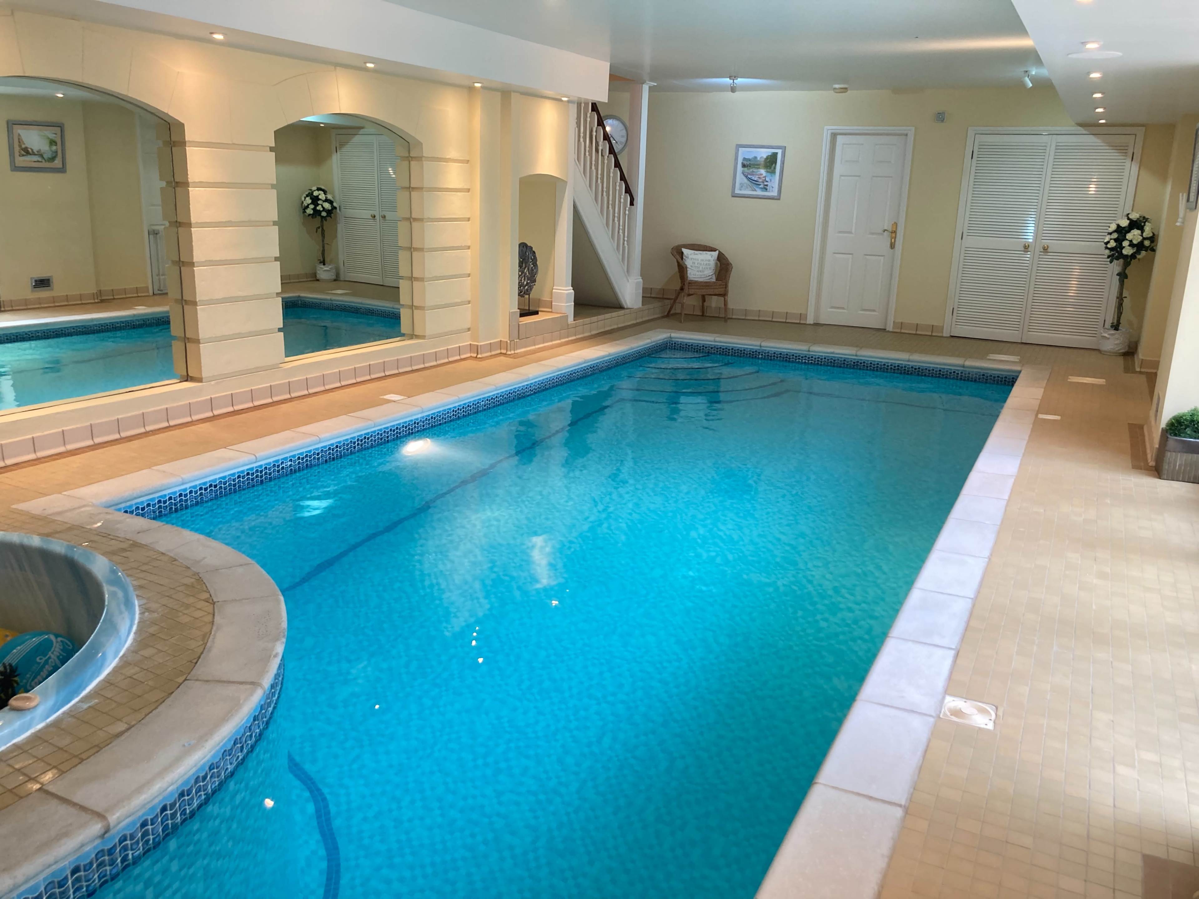 An indoor swimming pool area with blue water, surrounded by tiled walls and a jacuzzi on one side.