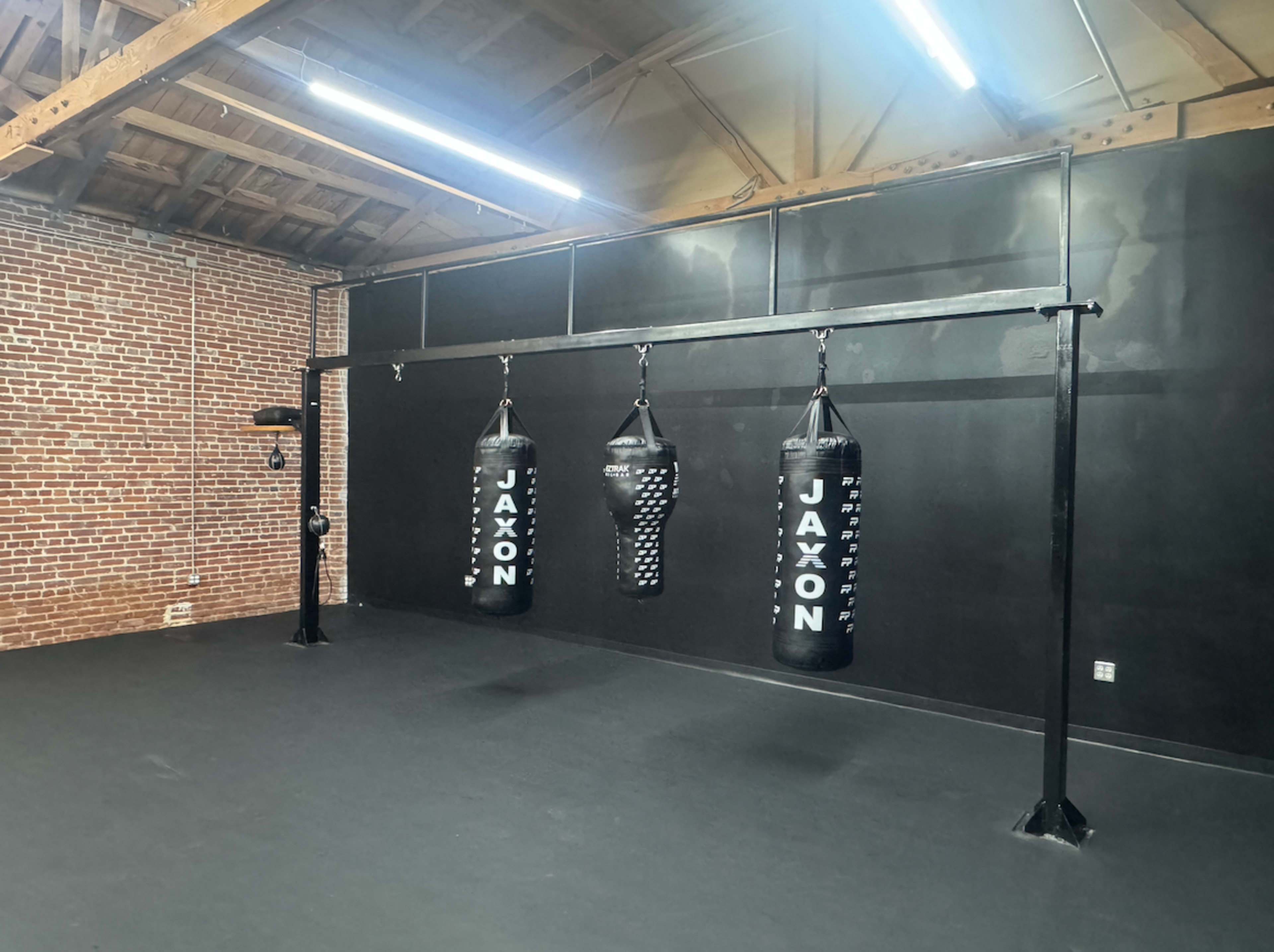 The image shows a gym or training space featuring three hanging black punching bags on a metal frame against a black wall with exposed brick.