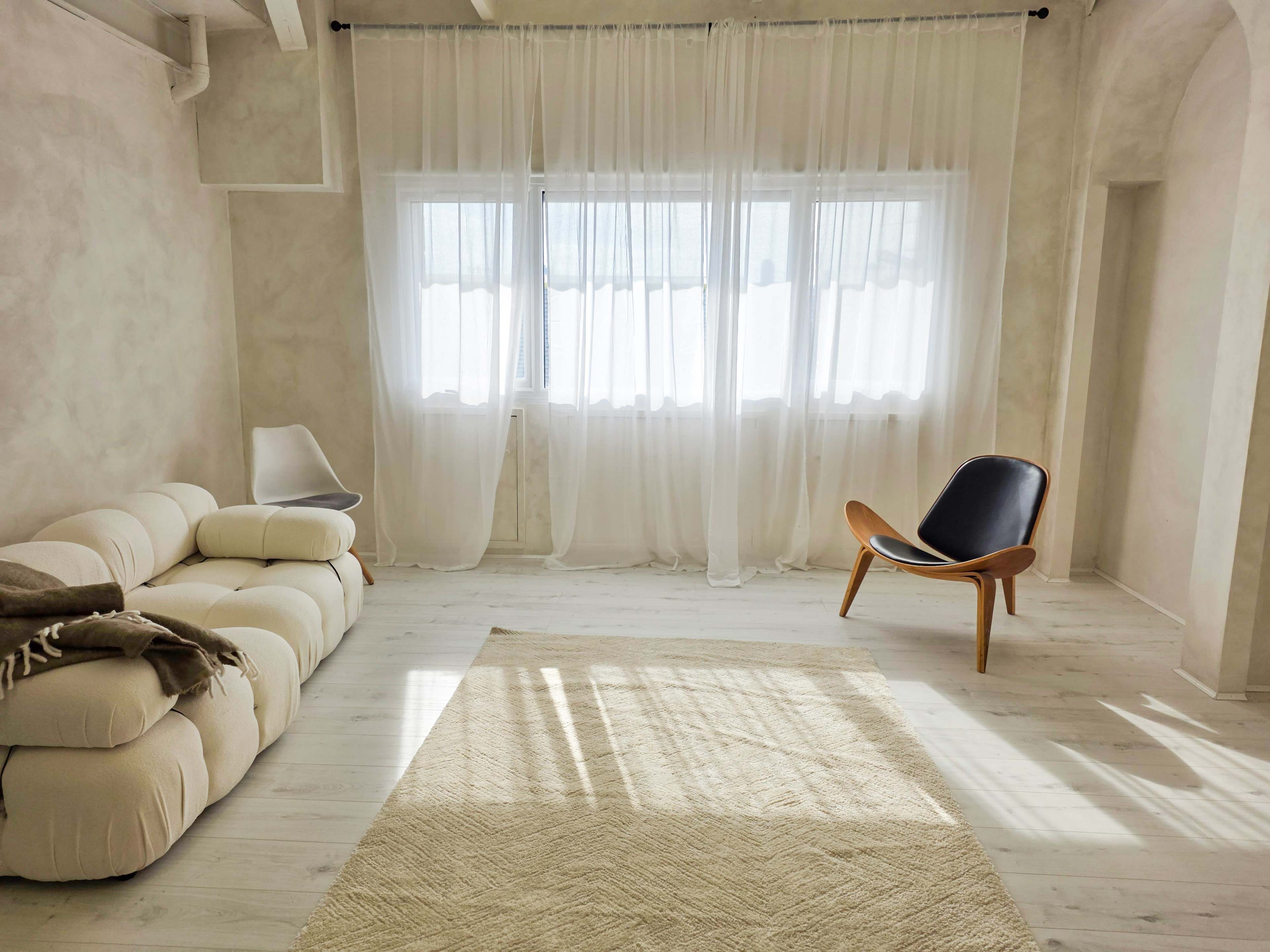 A bright, minimalist living room features a plush white sofa, a dark wooden chair, and large windows draped with sheer curtains.