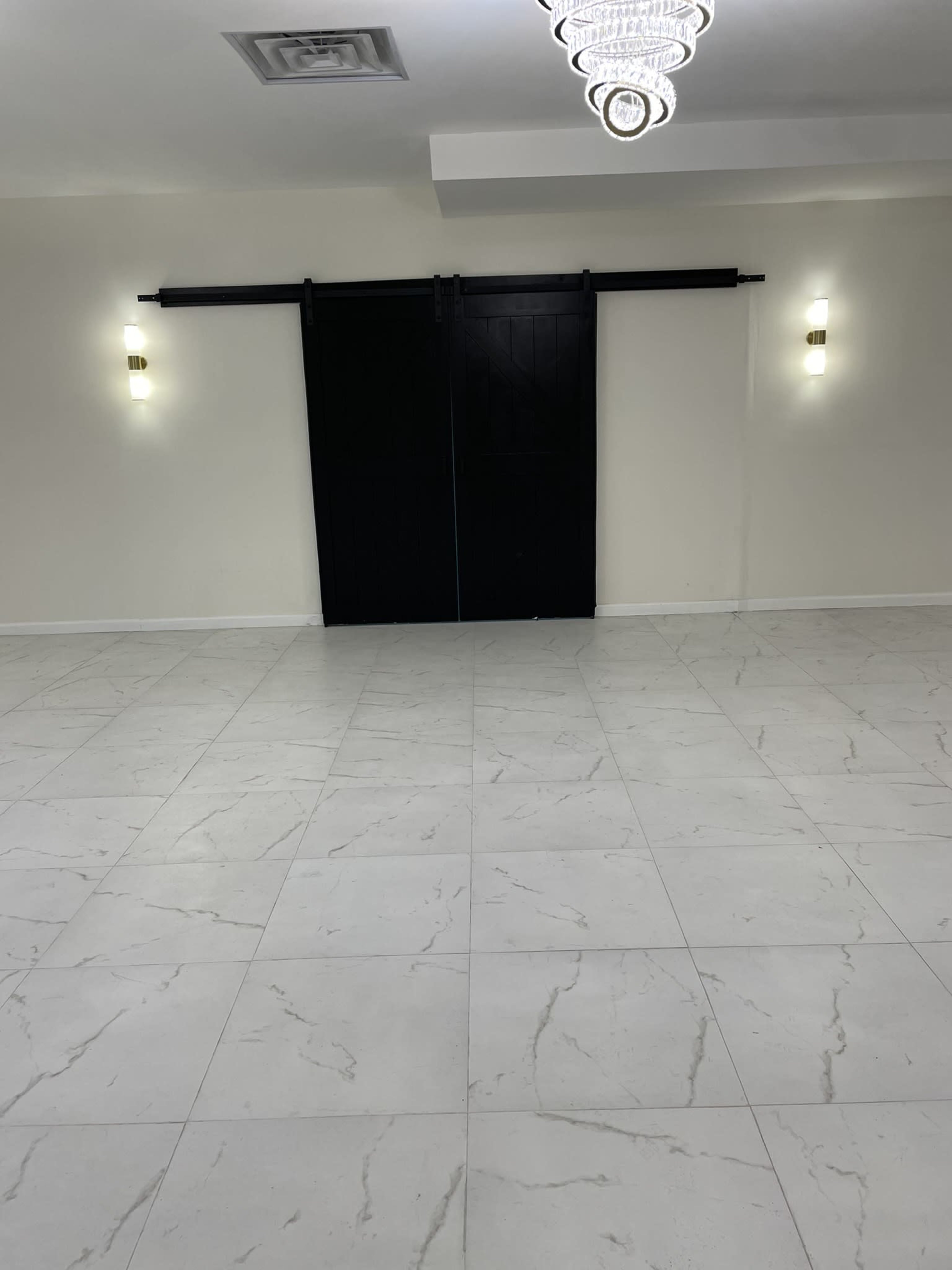 The image shows a spacious room with a tiled floor and black sliding doors at one end, illuminated by two wall-mounted lights and a chandelier above.