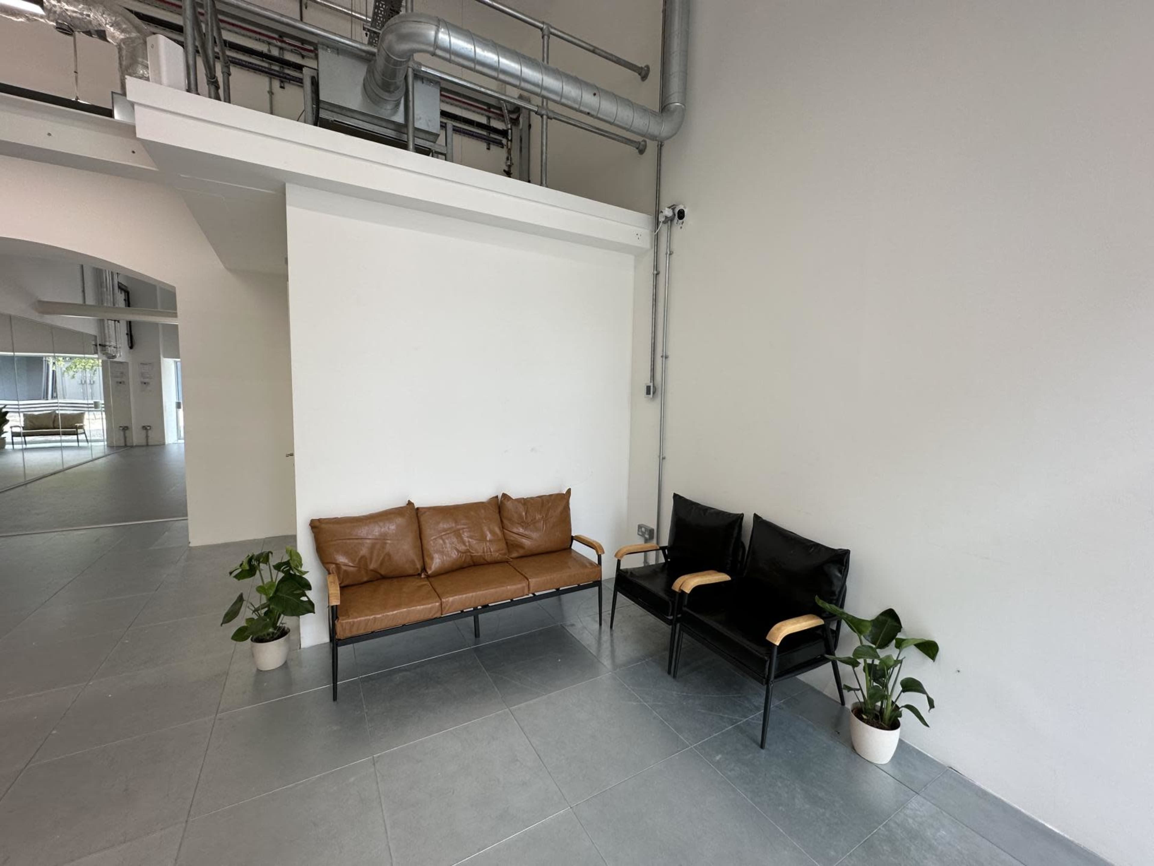 The image shows a small waiting area with a brown leather couch and two black chairs, accompanied by potted plants on the floor.