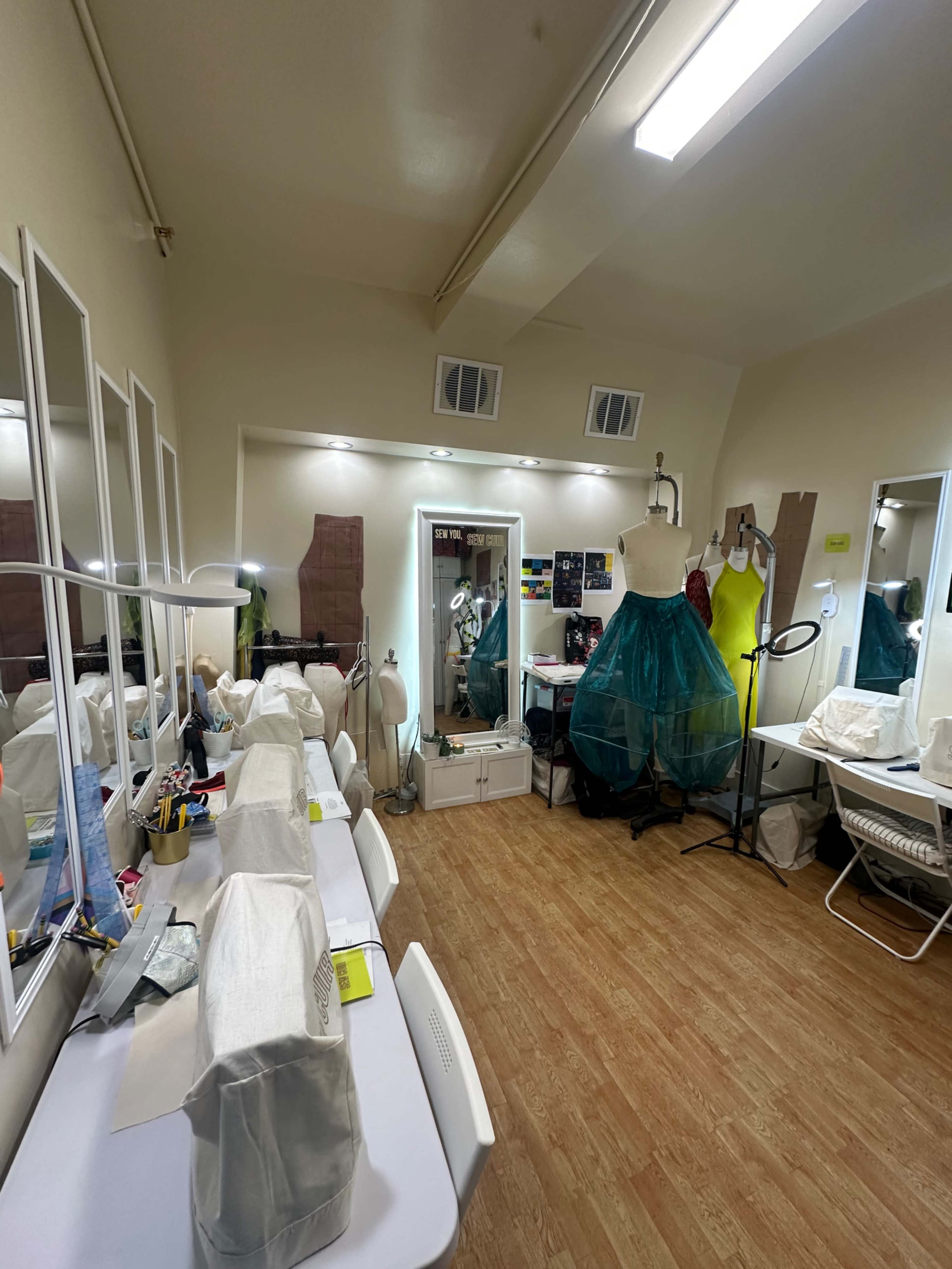 The image depicts a sewing studio featuring multiple workstations, a dress form, mirrors, and various sewing supplies scattered throughout the space.