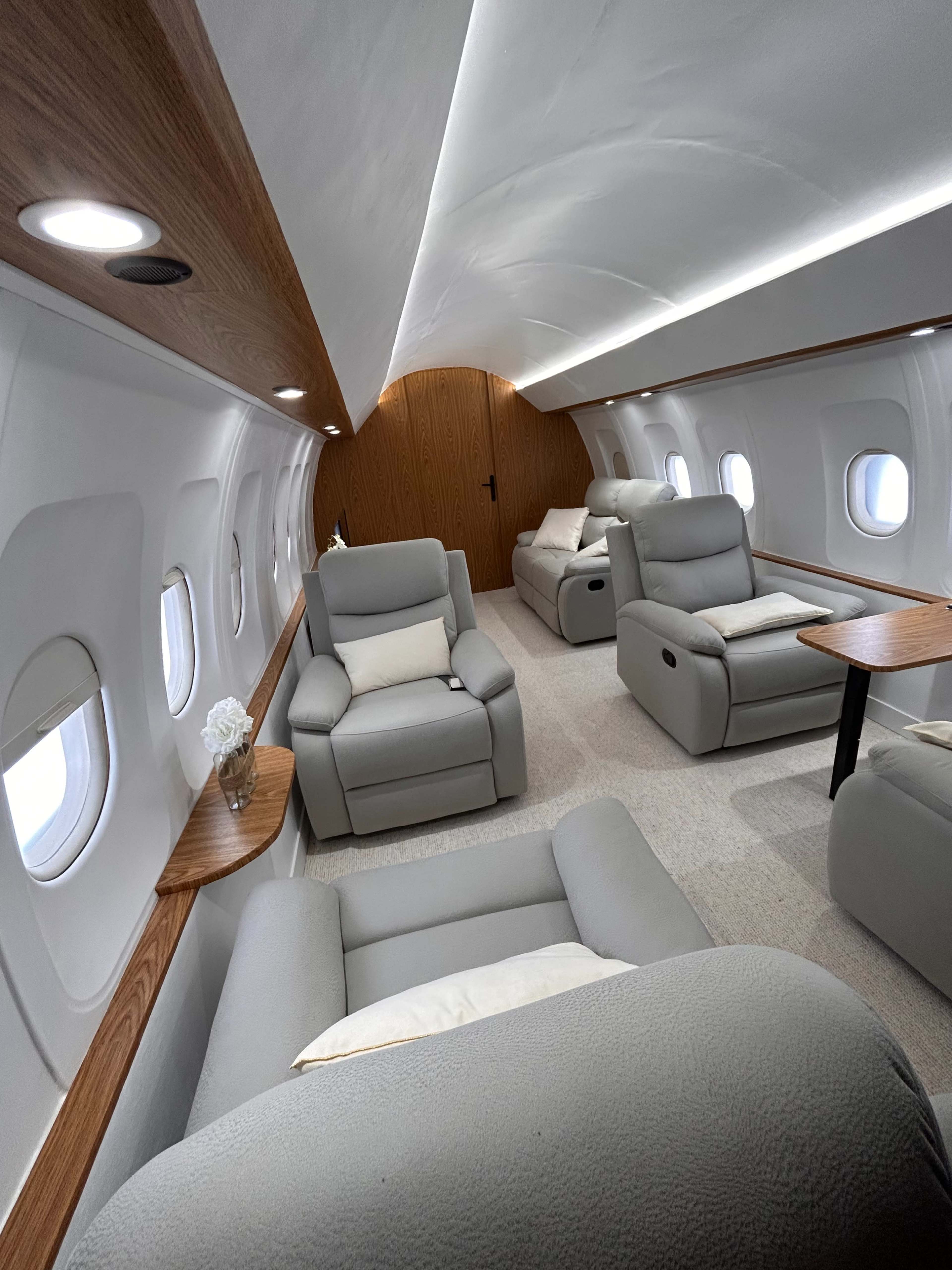 The interior of a private jet features several gray reclining chairs arranged around wooden tables, with soft lighting and small windows.