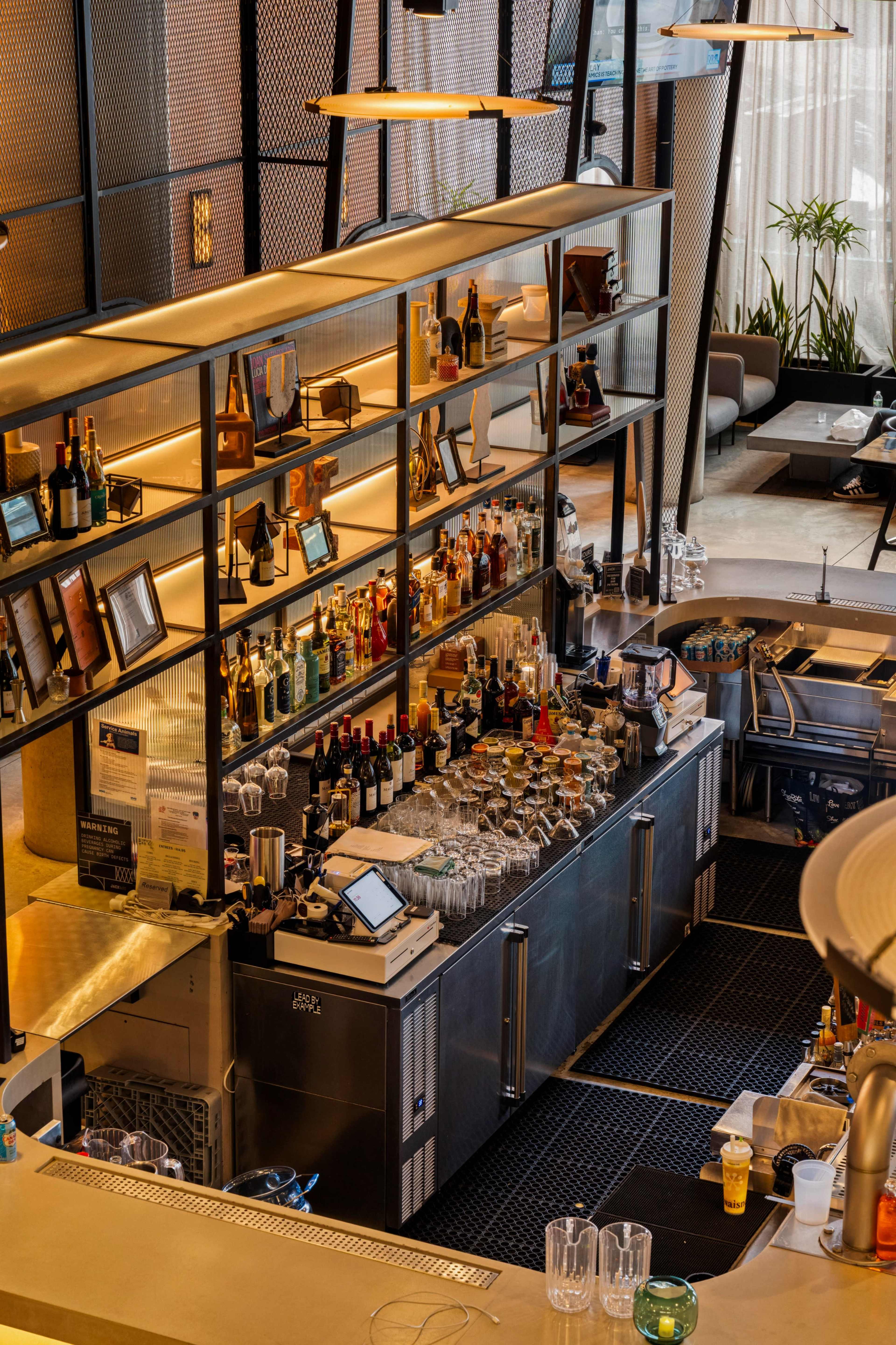 The image shows a modern bar with an illuminated display of various alcoholic beverages arranged neatly on shelves behind a sleek counter.