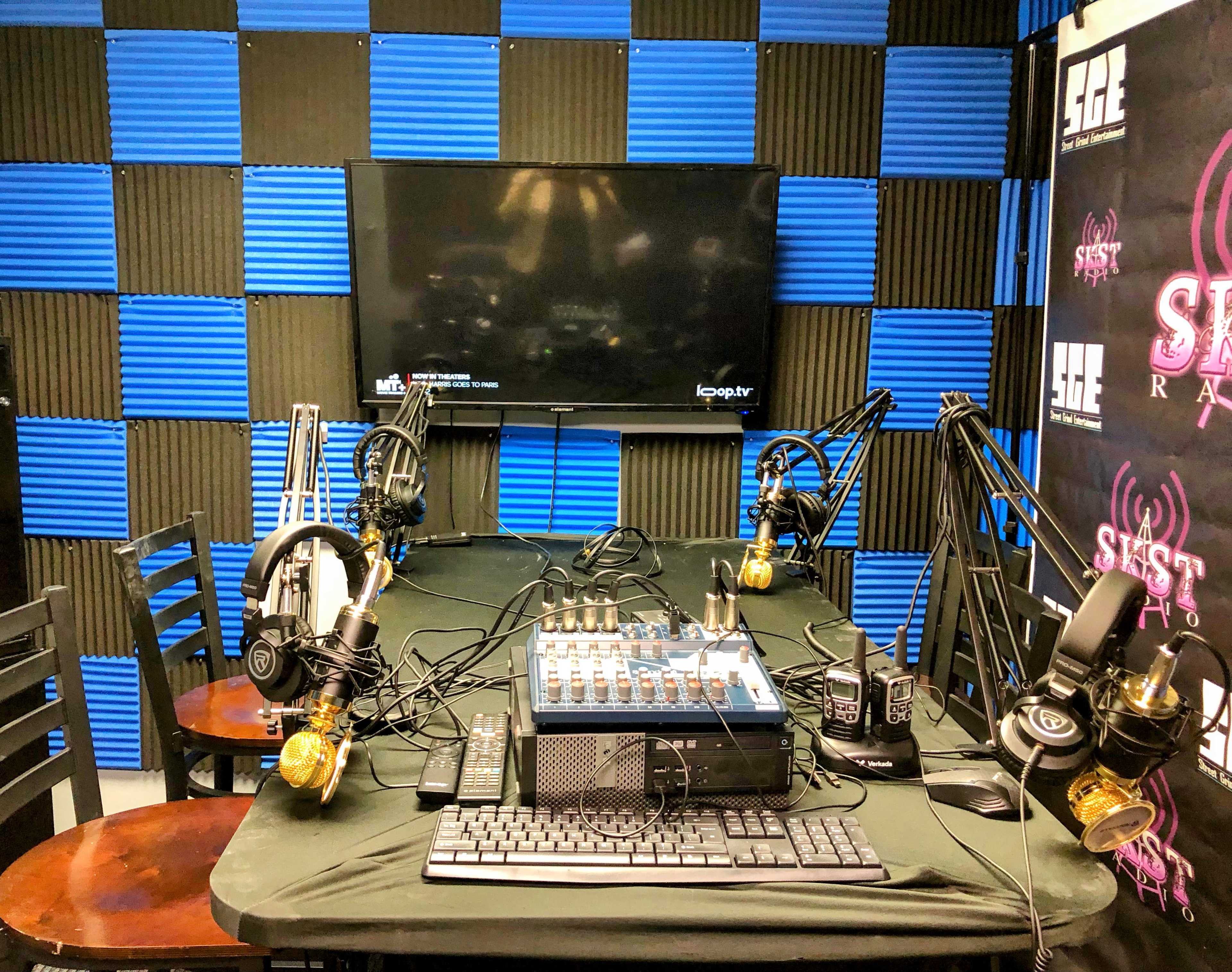 A radio studio equipped with microphones, headphones, and a mixing console, set against a backdrop of blue and black acoustic panels.