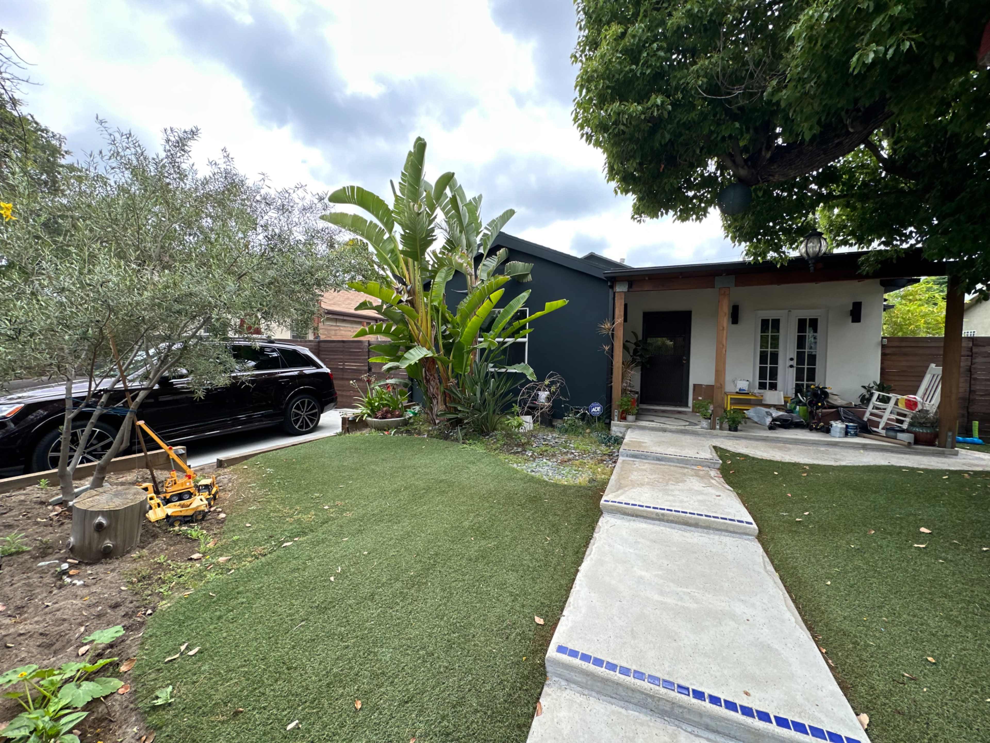 The image shows a modern house with a well-maintained front garden featuring artificial grass, a pathway leading to the entrance, and a parked black SUV on the side.