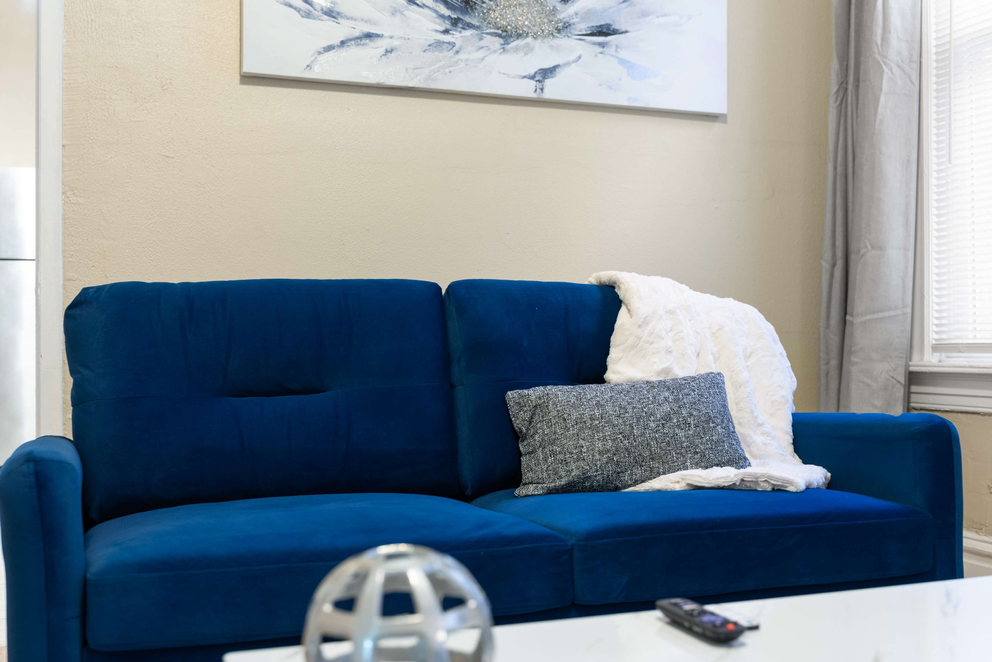 A blue couch in a living room is adorned with a gray pillow and a white throw blanket, next to a coffee table with a decorative globe and a remote control.
