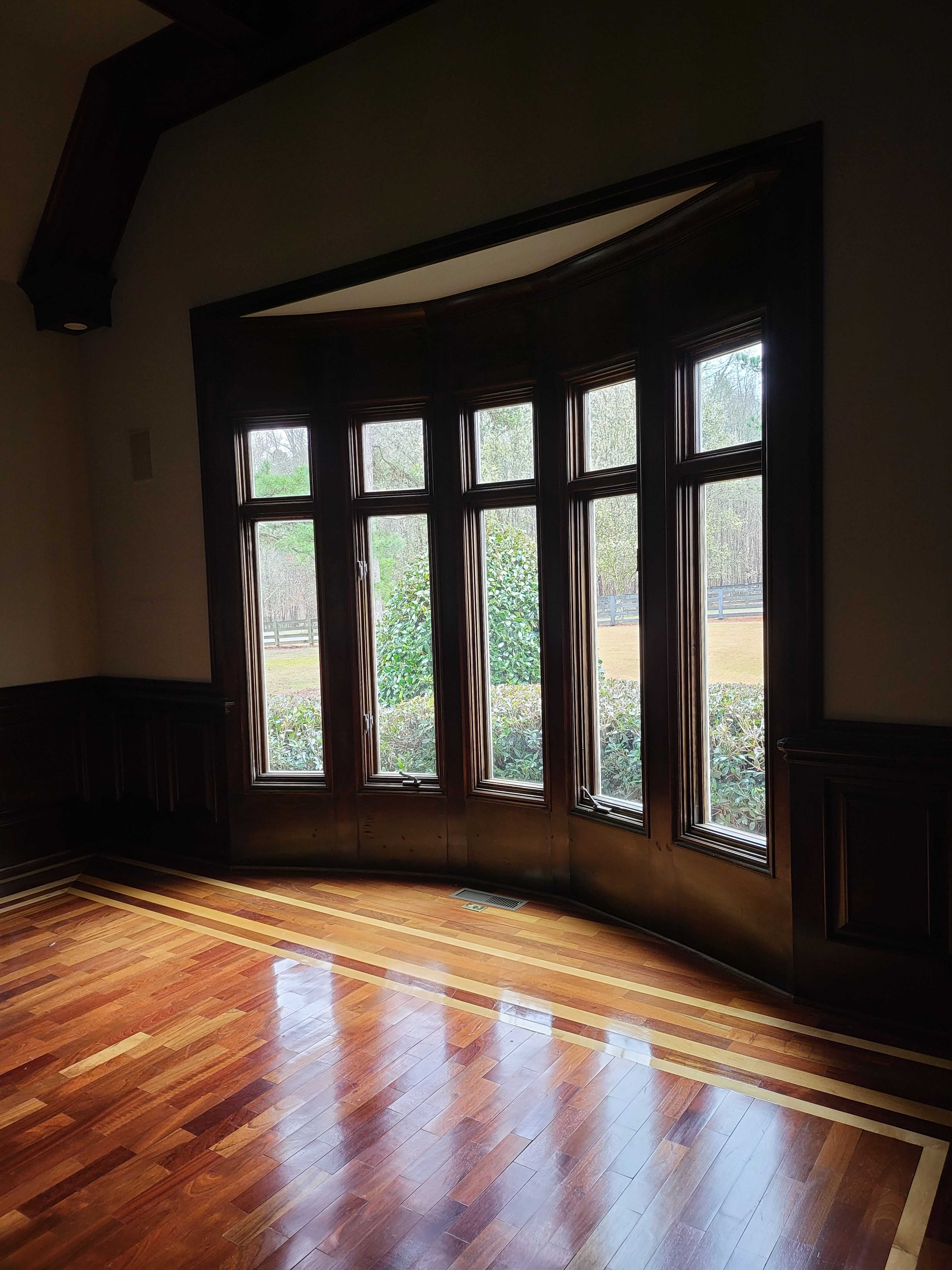 A bay window with multiple panes overlooks a landscaped area from a room with hardwood flooring.