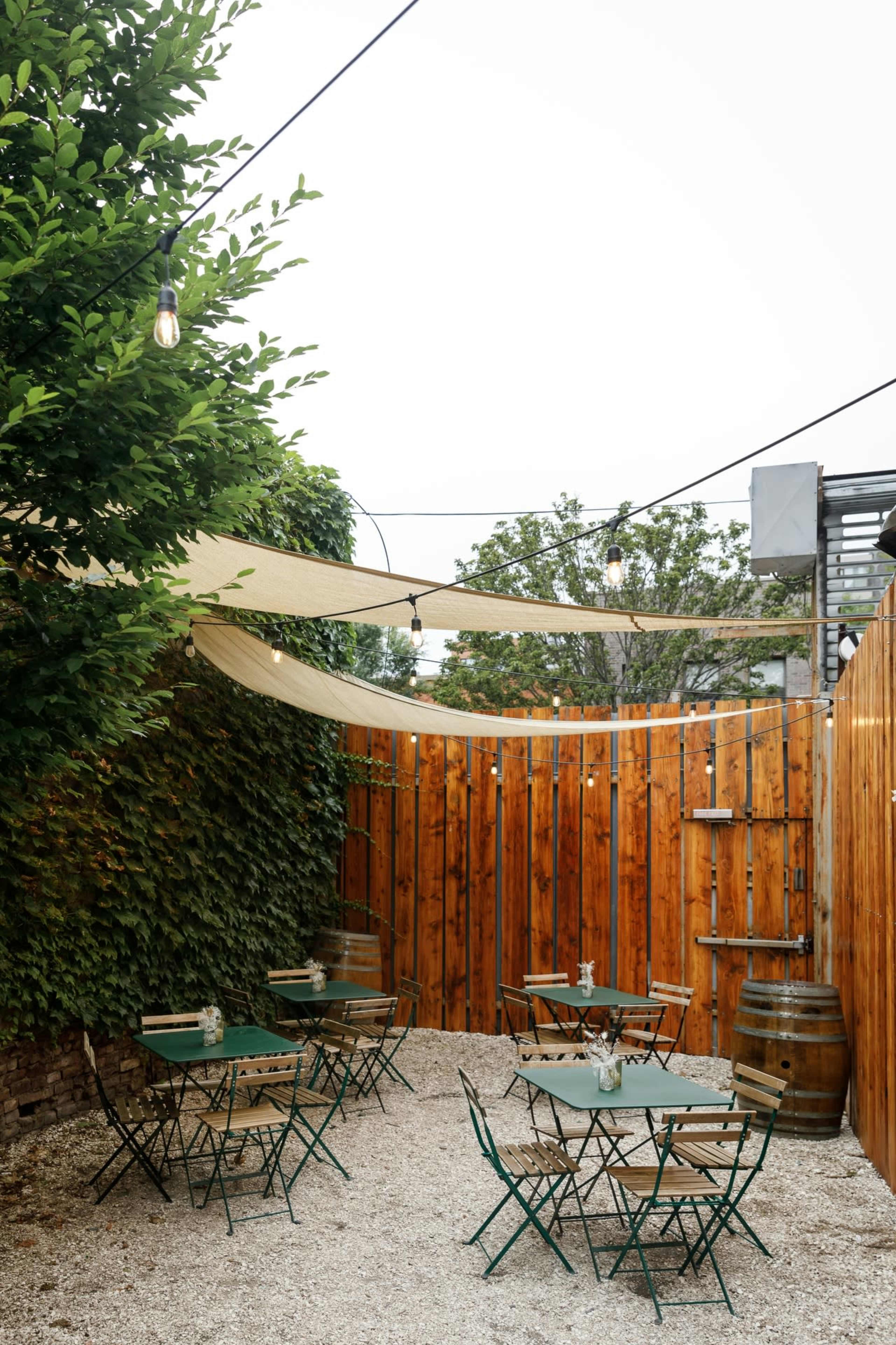 The image shows an outdoor café area with wooden fencing, green tables and chairs, and string lights overhead, surrounded by greenery.
