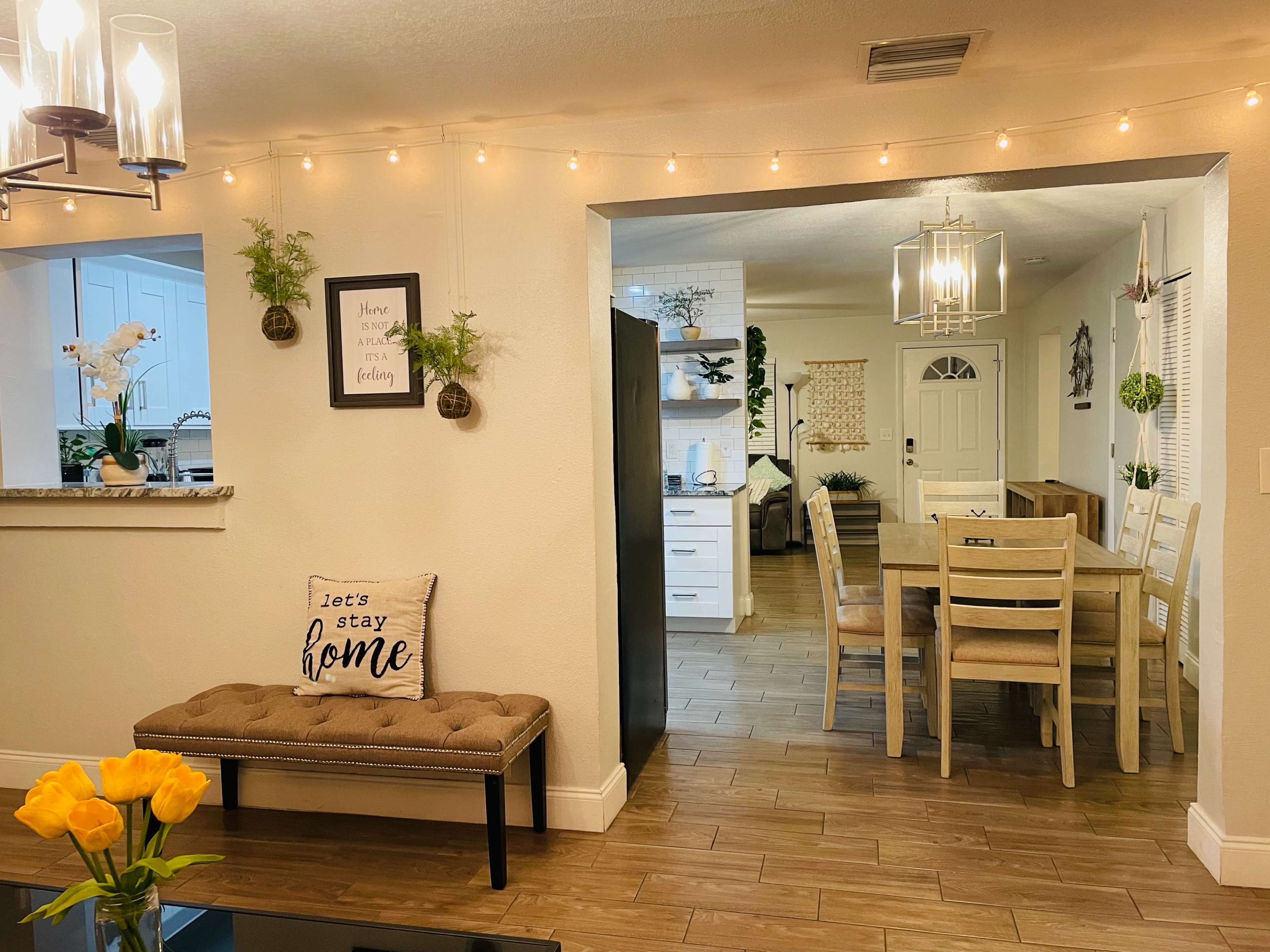 A cozy living area features a plush bench with a "let's stay home" pillow, leading to a dining space with a wooden table and chairs, adorned with warm string lights.