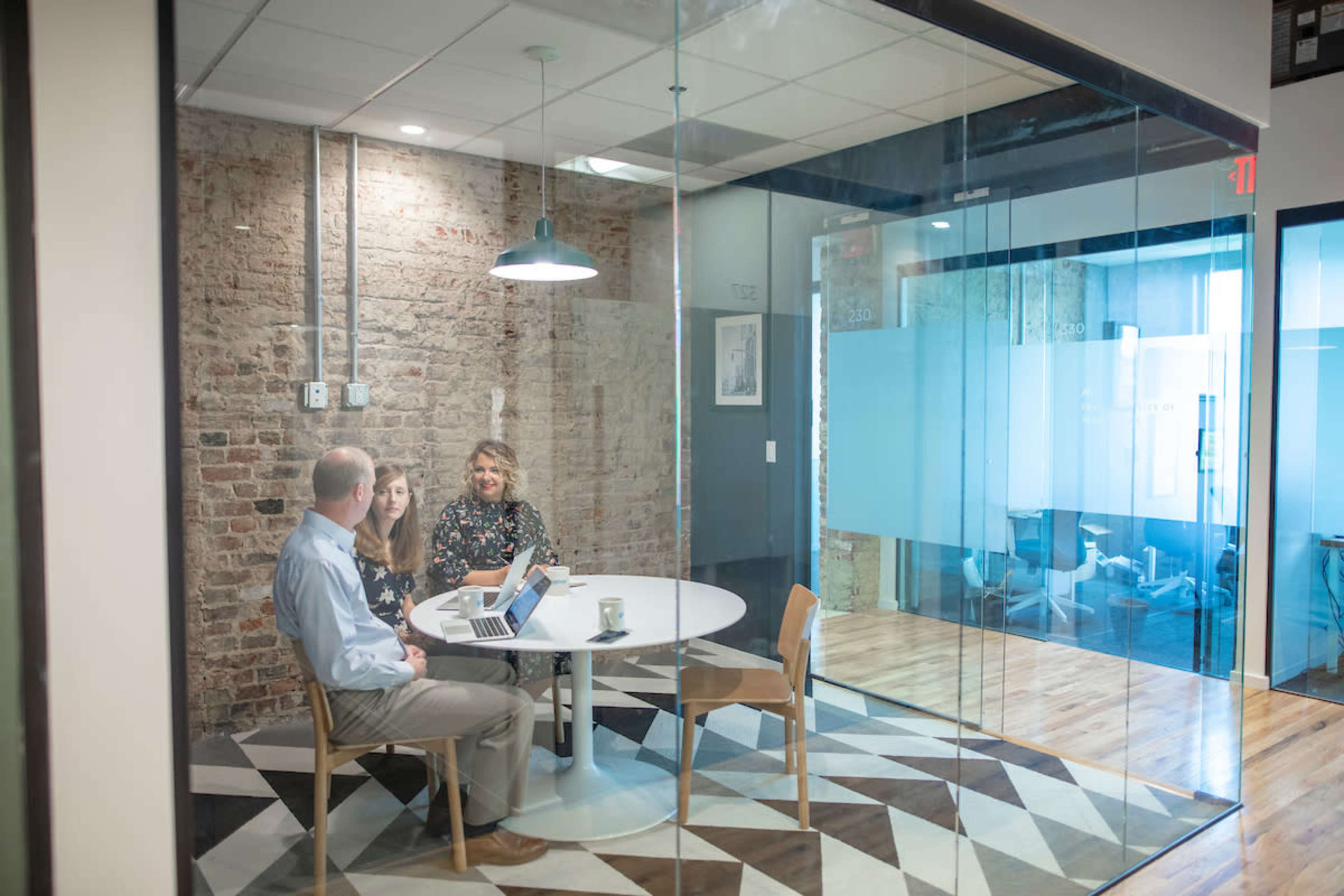Three people are seated at a round table in a glass-walled conference room with exposed brick, discussing while using a laptop.