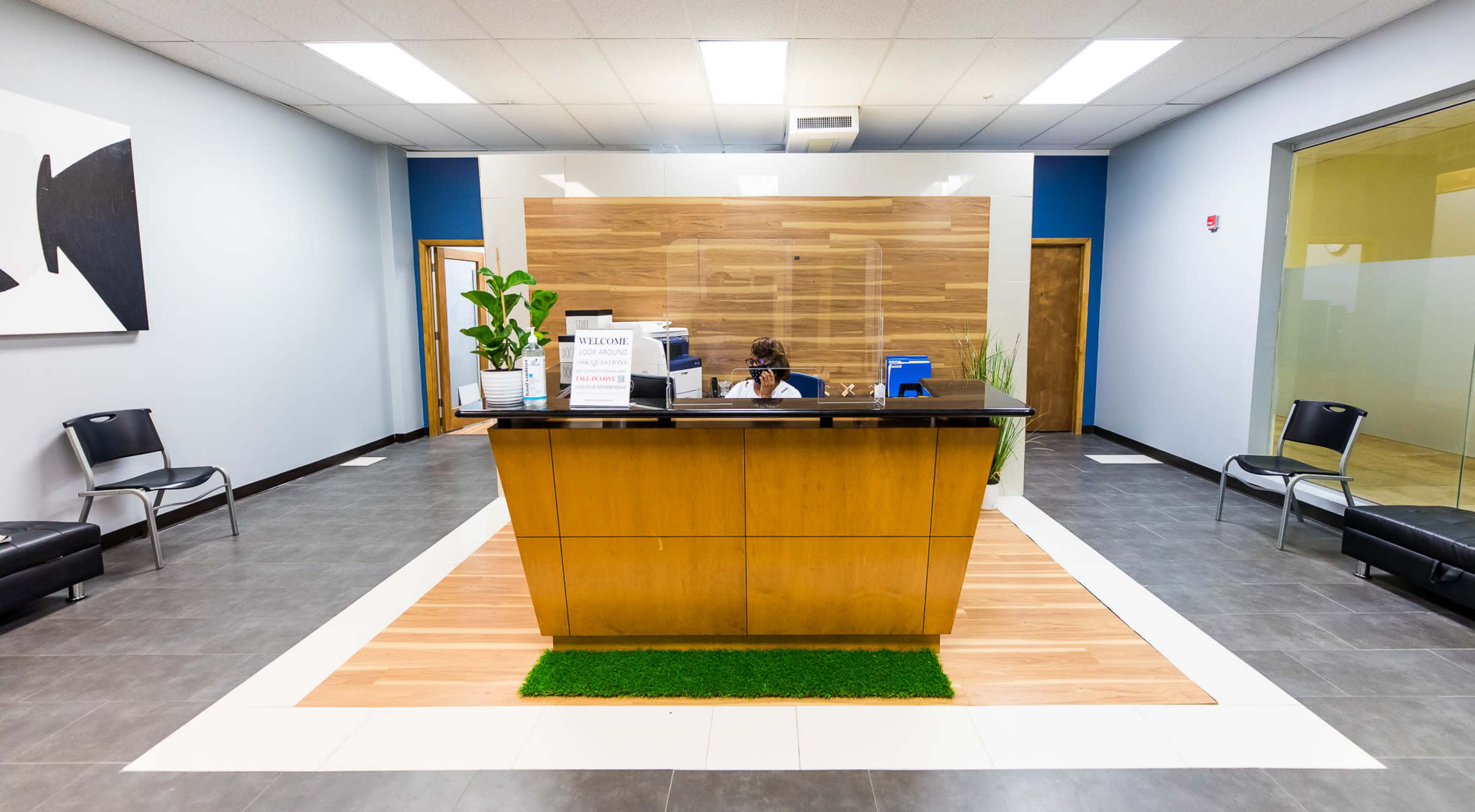 A modern reception area featuring a wooden reception desk, indoor plants, and seating on either side with gray tile flooring.