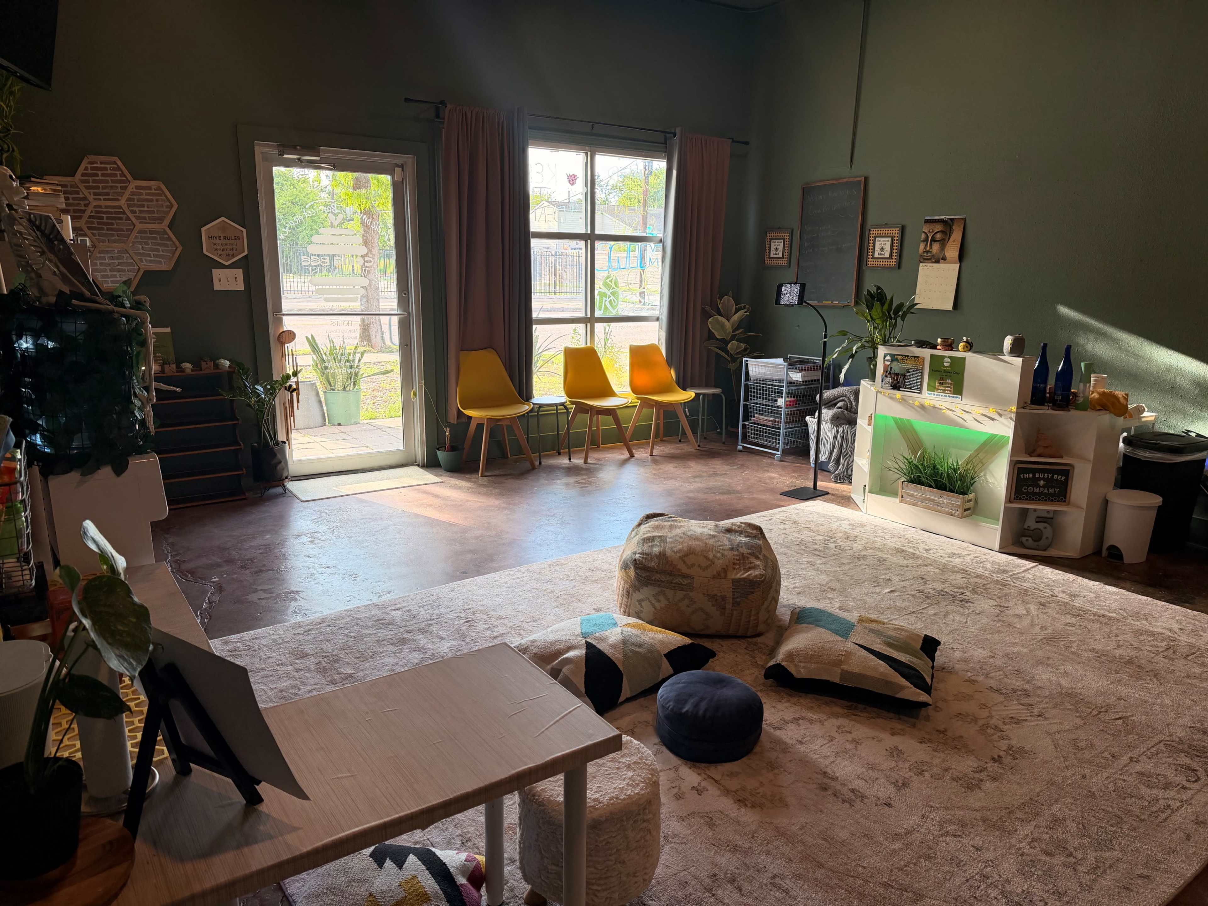 The image shows a well-lit room with a seating area featuring yellow chairs, a cozy rug, and decorative plants, along with a small table and storage units.