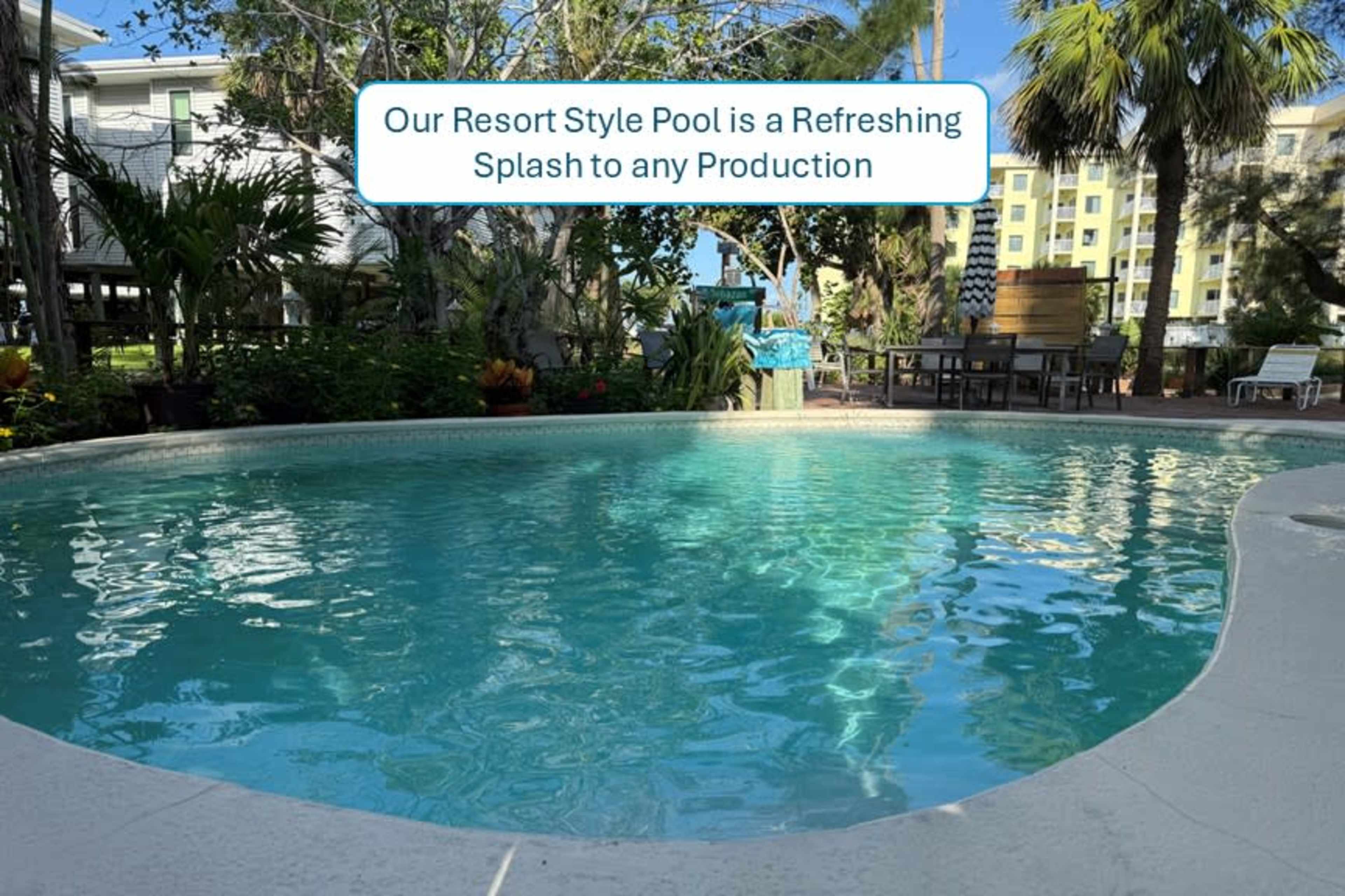 The image shows a clear, inviting pool surrounded by tropical plants and lounge chairs in a resort-like setting.