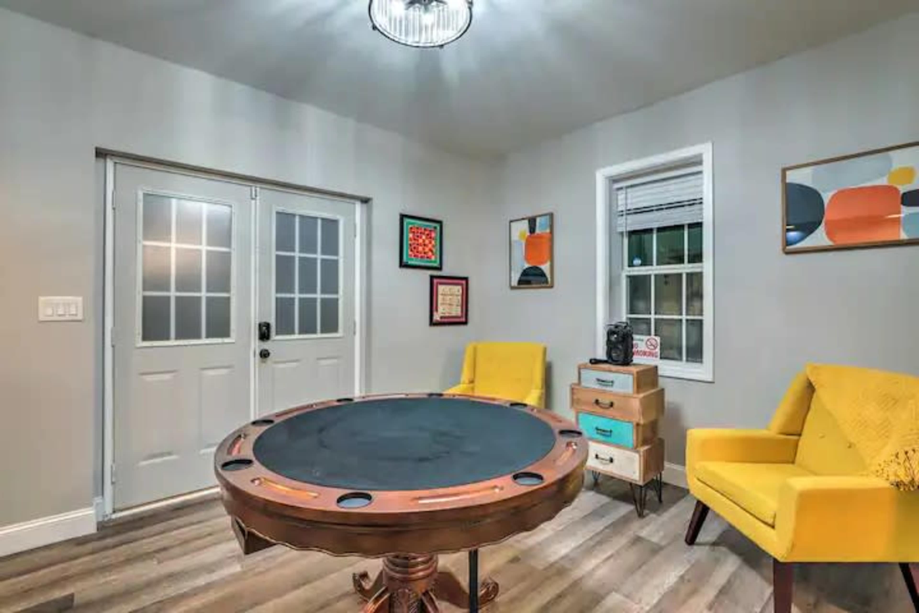 A room featuring a round poker table at the center, flanked by two yellow chairs, with double doors leading outside and boxes stacked against the wall.