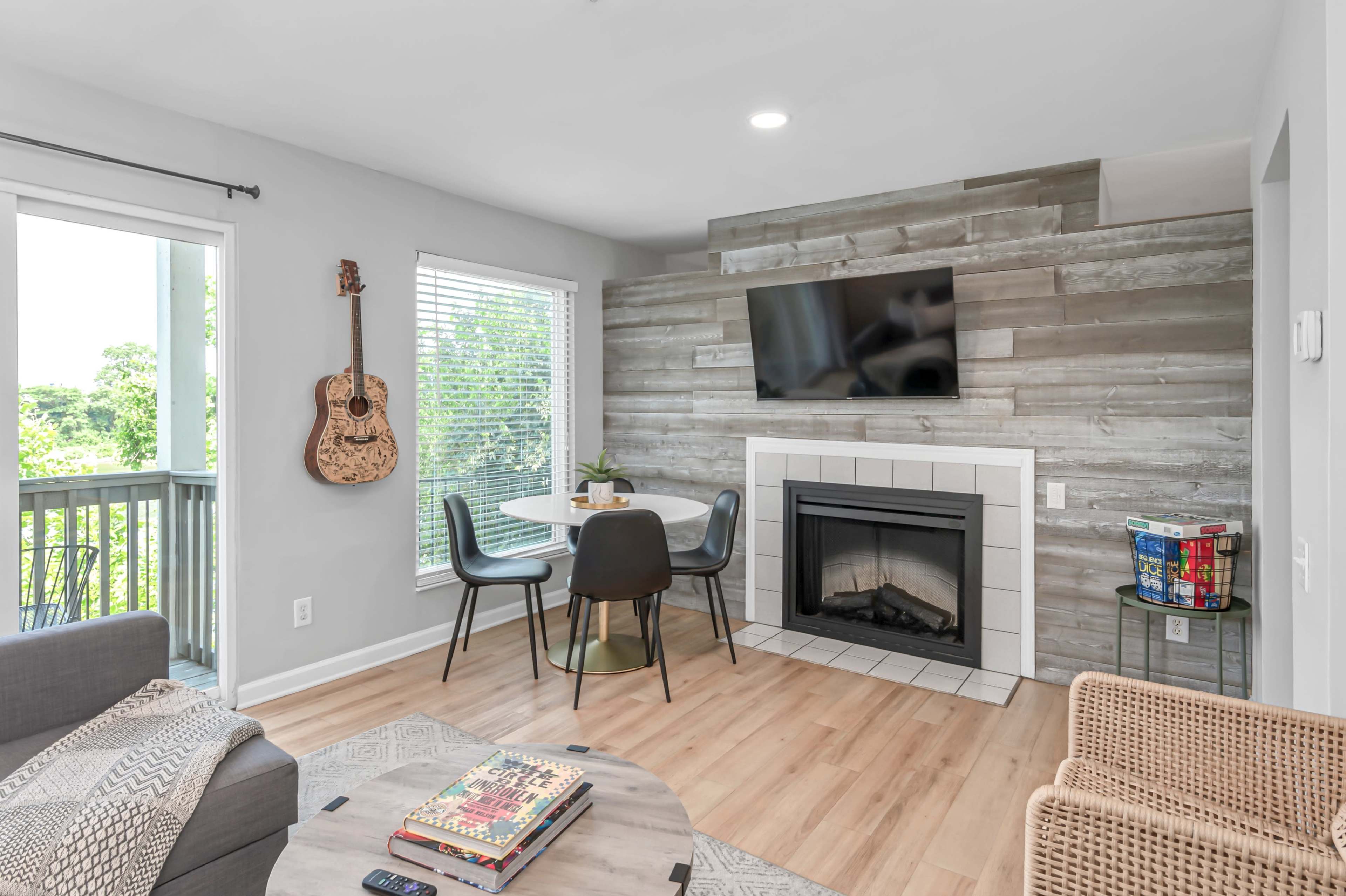 A living area features a wall-mounted television above a fireplace, surrounded by a dining table and chairs, with a guitar and large windows in the background.
