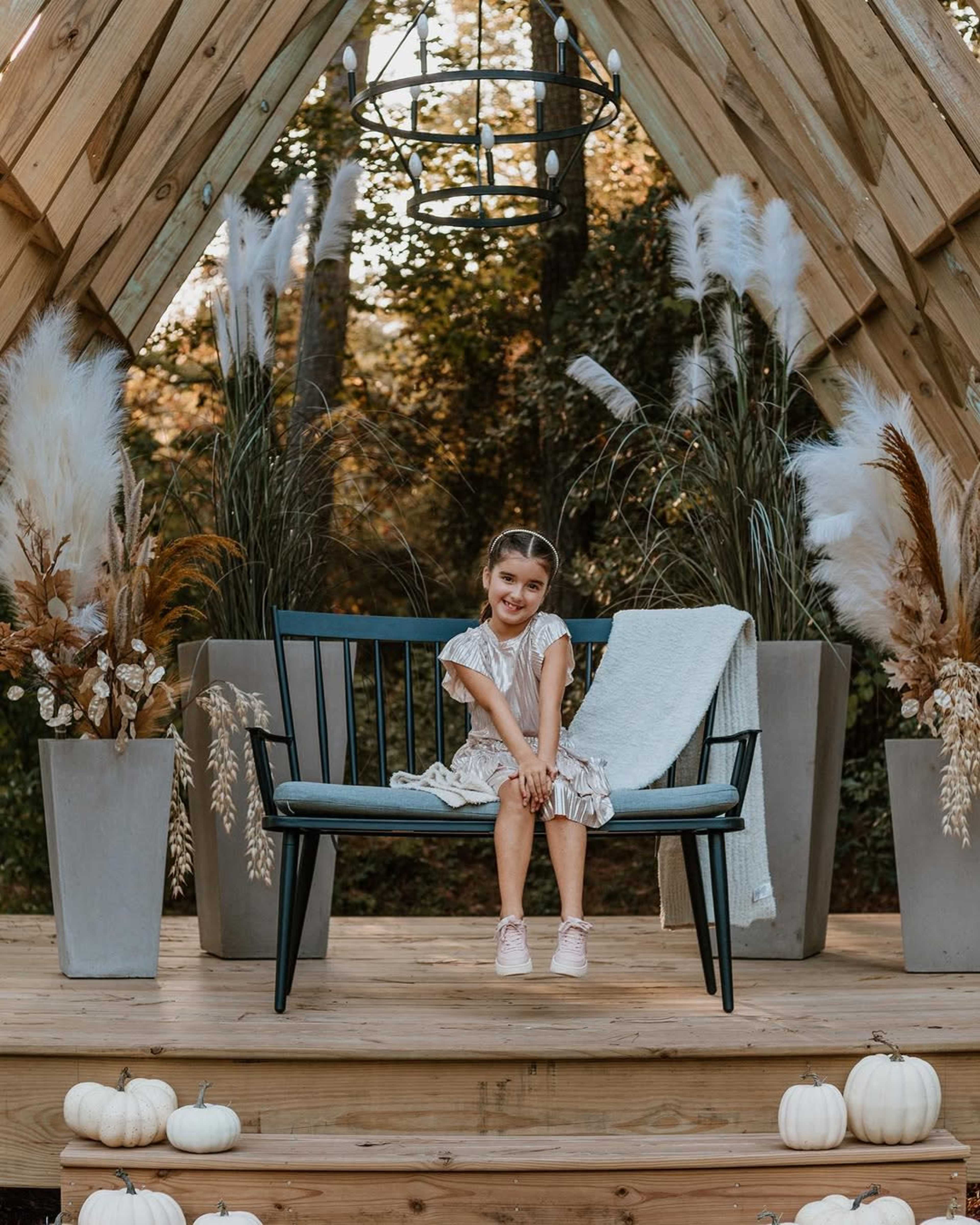 A young girl in a dress sits on a black bench surrounded by decorative plants and pumpkins under a wooden archway.