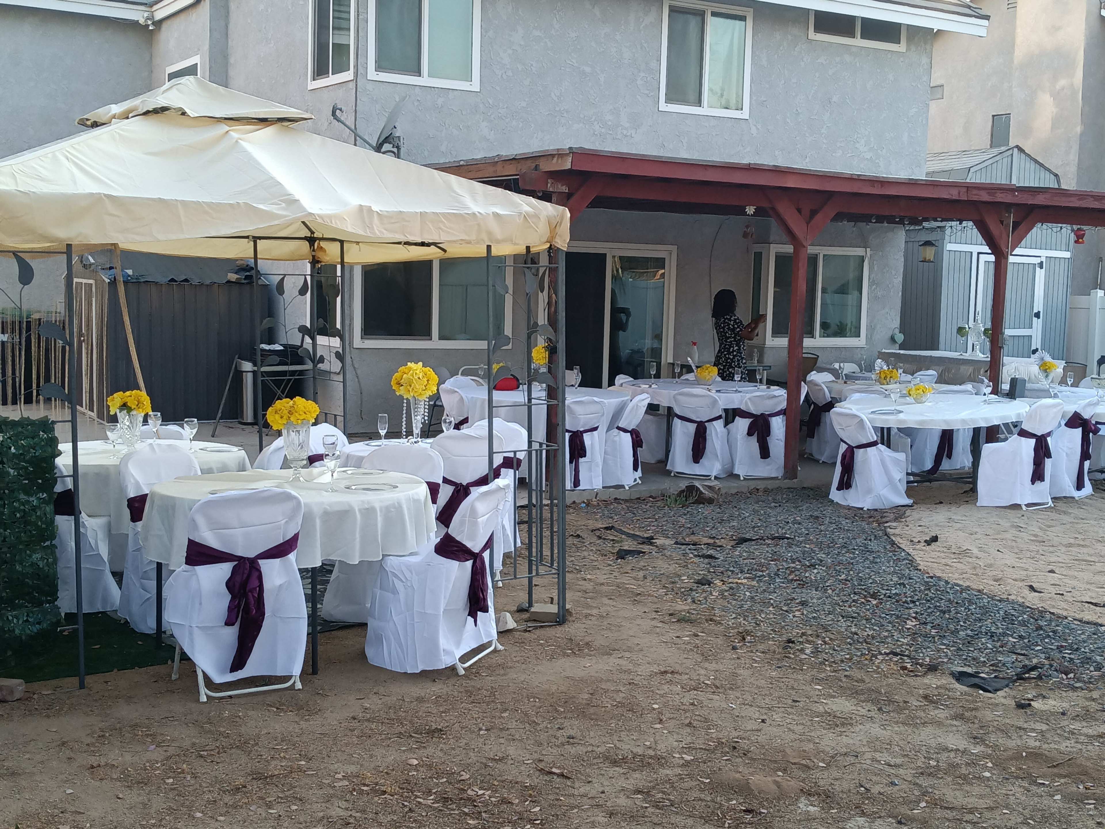 A tent is set up in a backyard with neatly arranged tables covered in white tablecloths and adorned with yellow flower centerpieces.