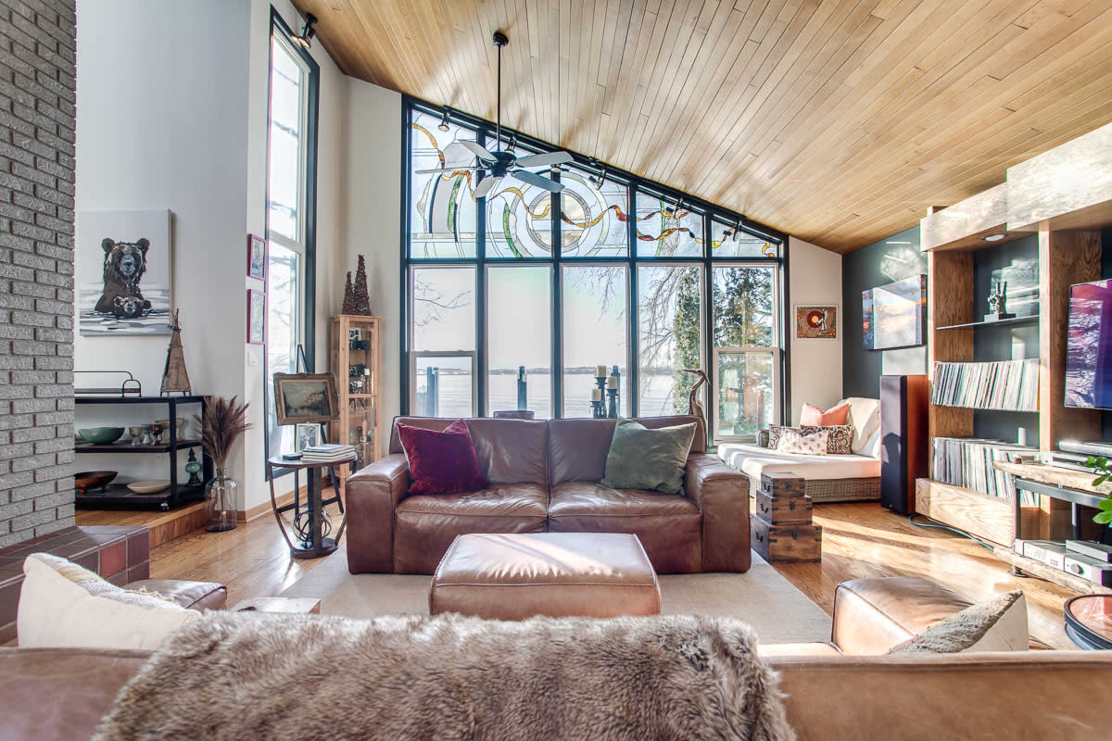 A spacious living room features large windows with a view of the water, wooden ceilings, and comfortable seating arrangements.