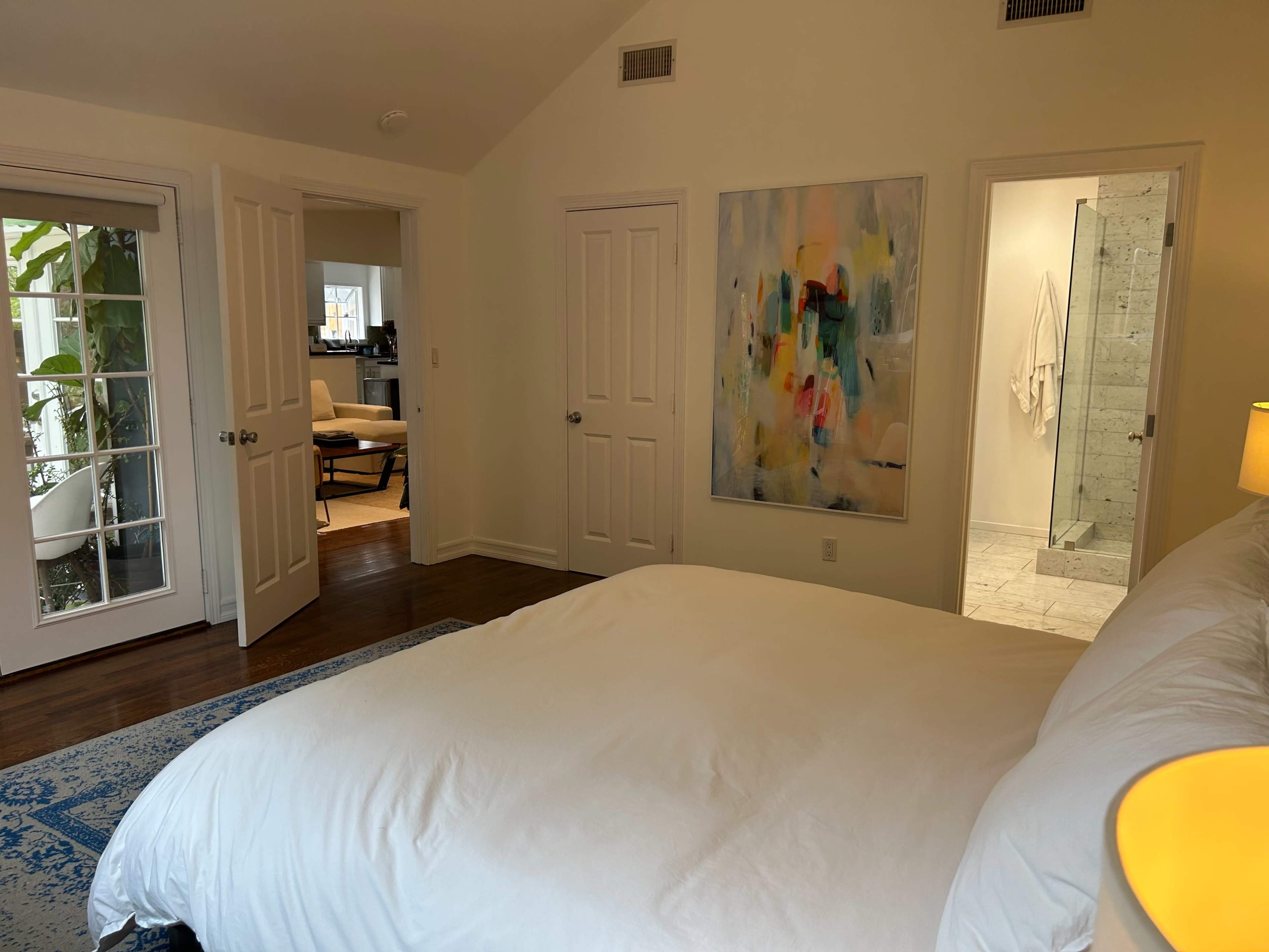 The image shows a bright bedroom with a large white bed, an abstract painting on the wall, and doors leading to another room and an outdoor area.