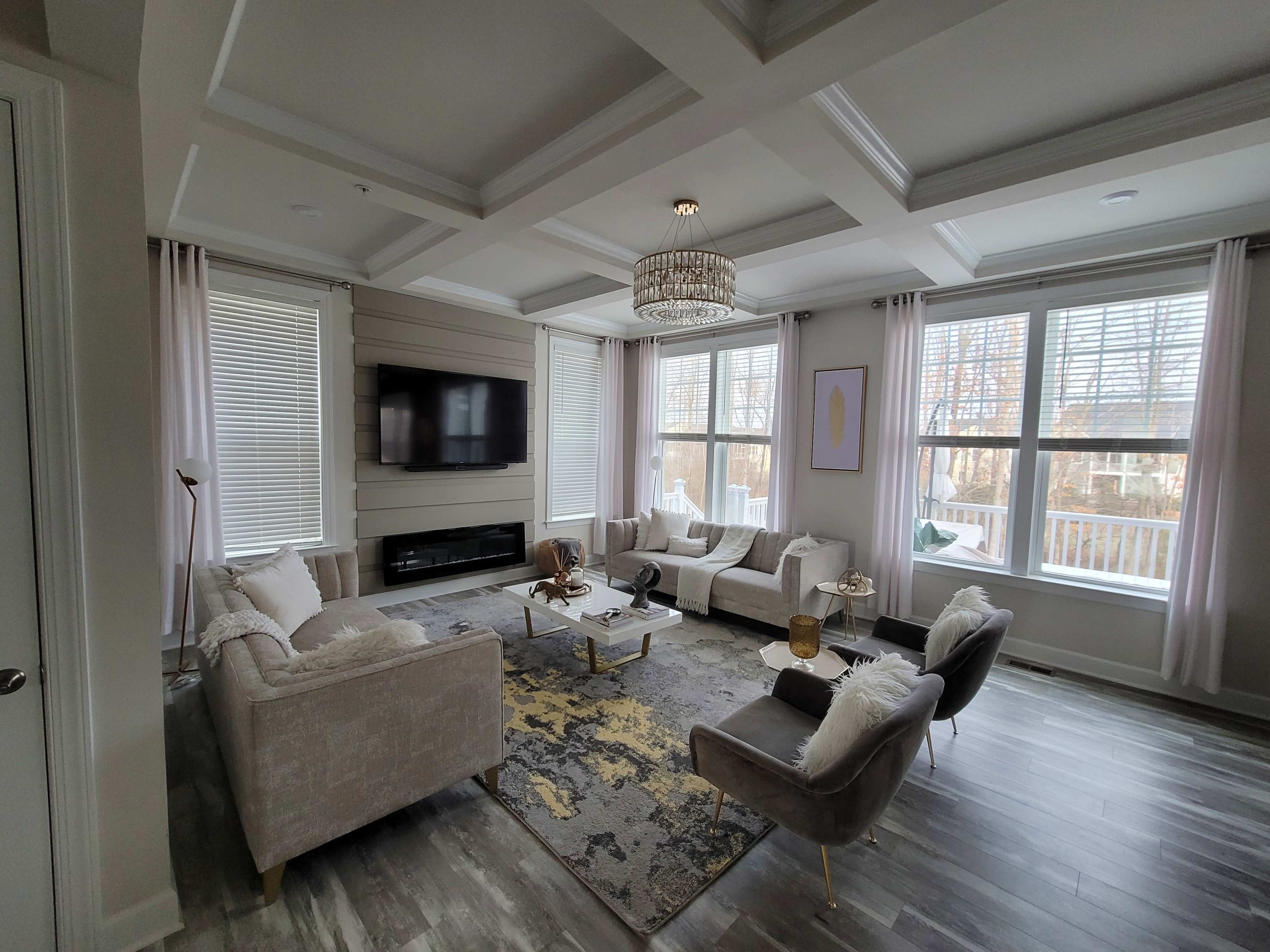 The room features a modern living area with a large sectional sofa, a coffee table, a wall-mounted TV, and large windows providing natural light.