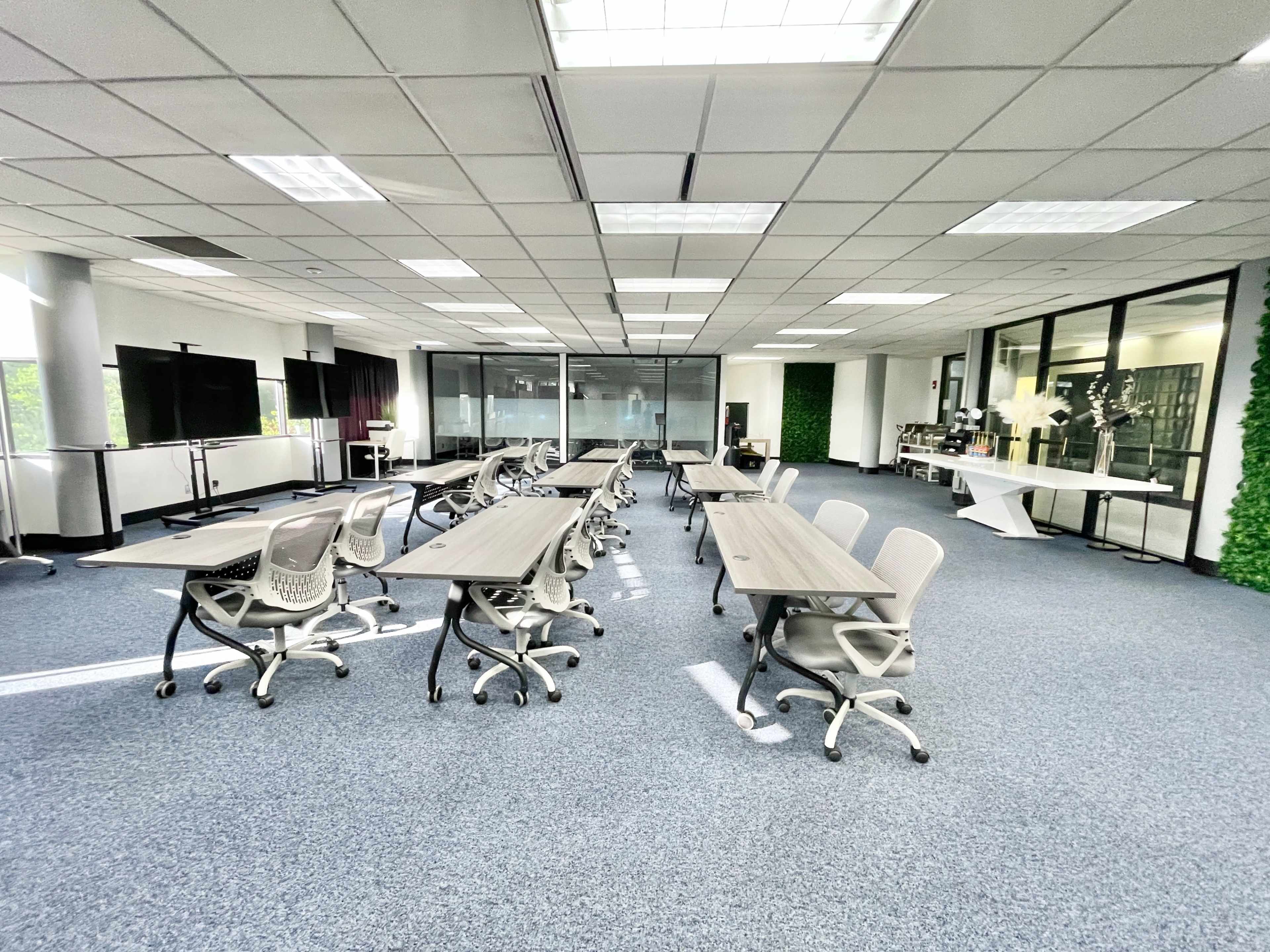 A spacious, well-lit office interior features several long tables arranged with ergonomic chairs, surrounded by glass walls and greenery accents.