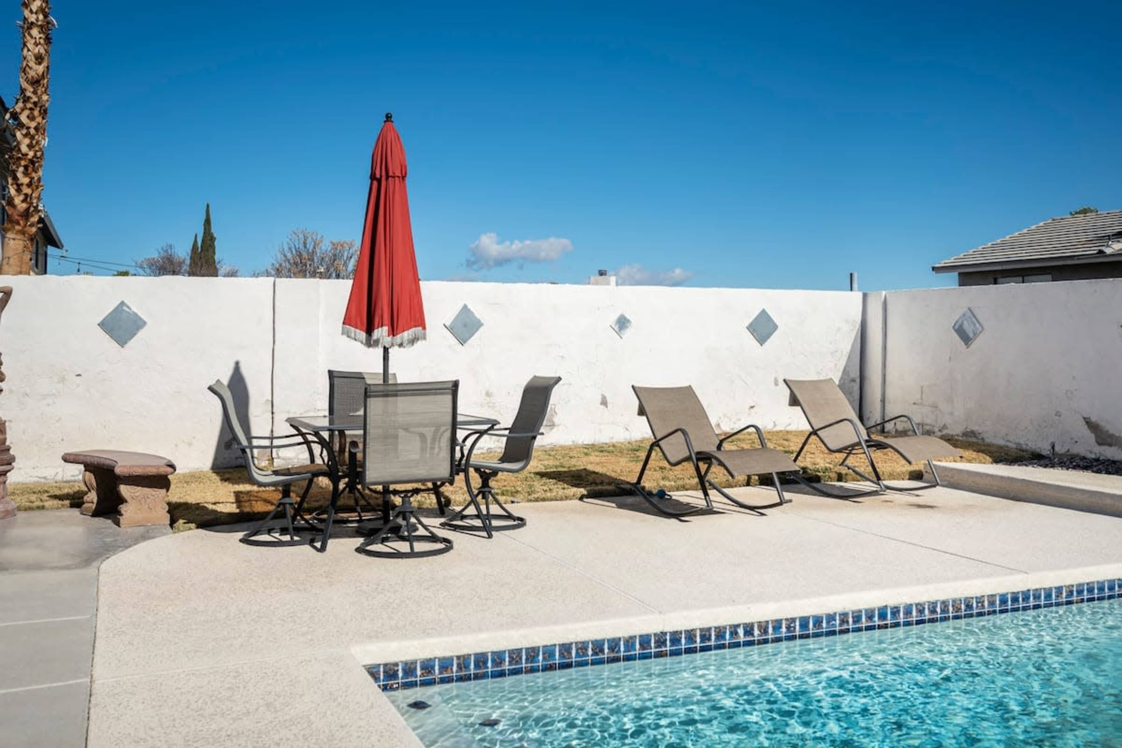 A patio area features a swimming pool, several lounge chairs, a table with chairs, and a red umbrella.