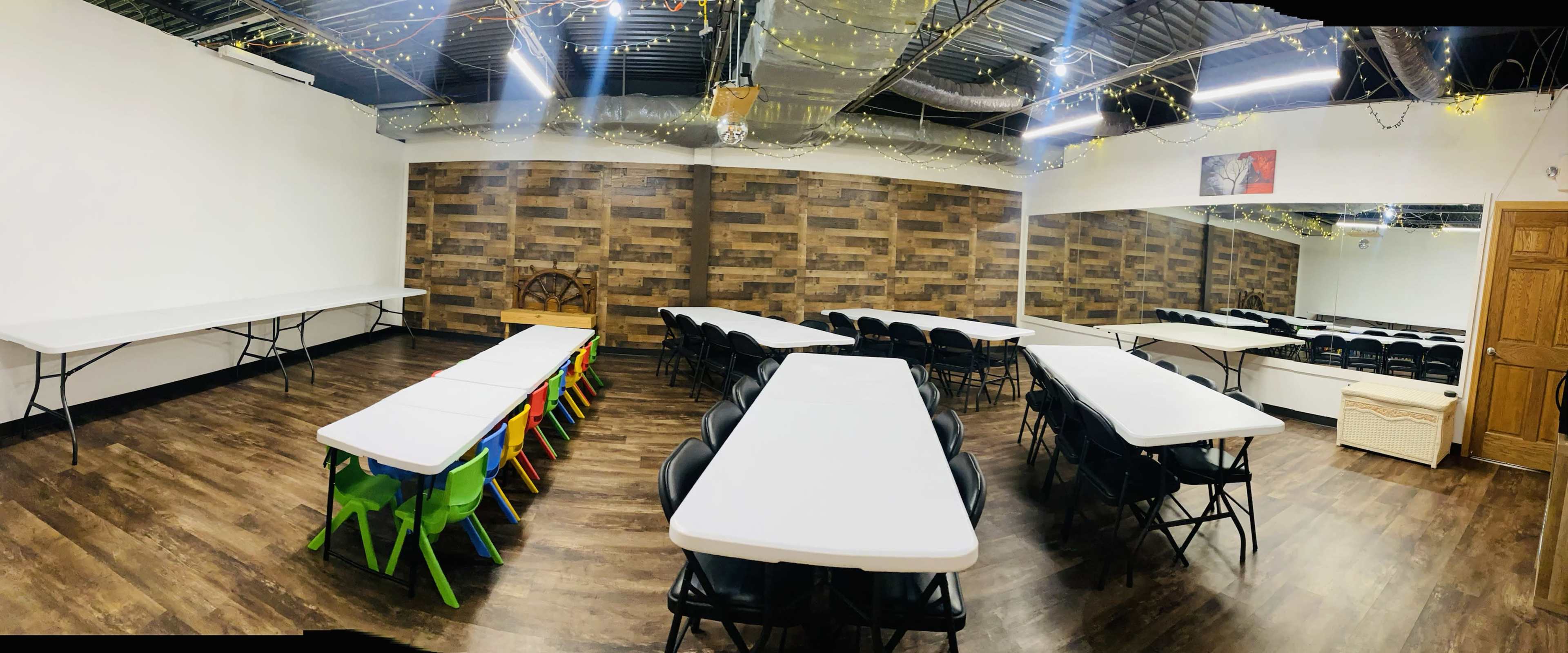 A spacious meeting room with multiple long tables arranged in a U-shape, black chairs, and wooden accent walls, illuminated by string lights.
