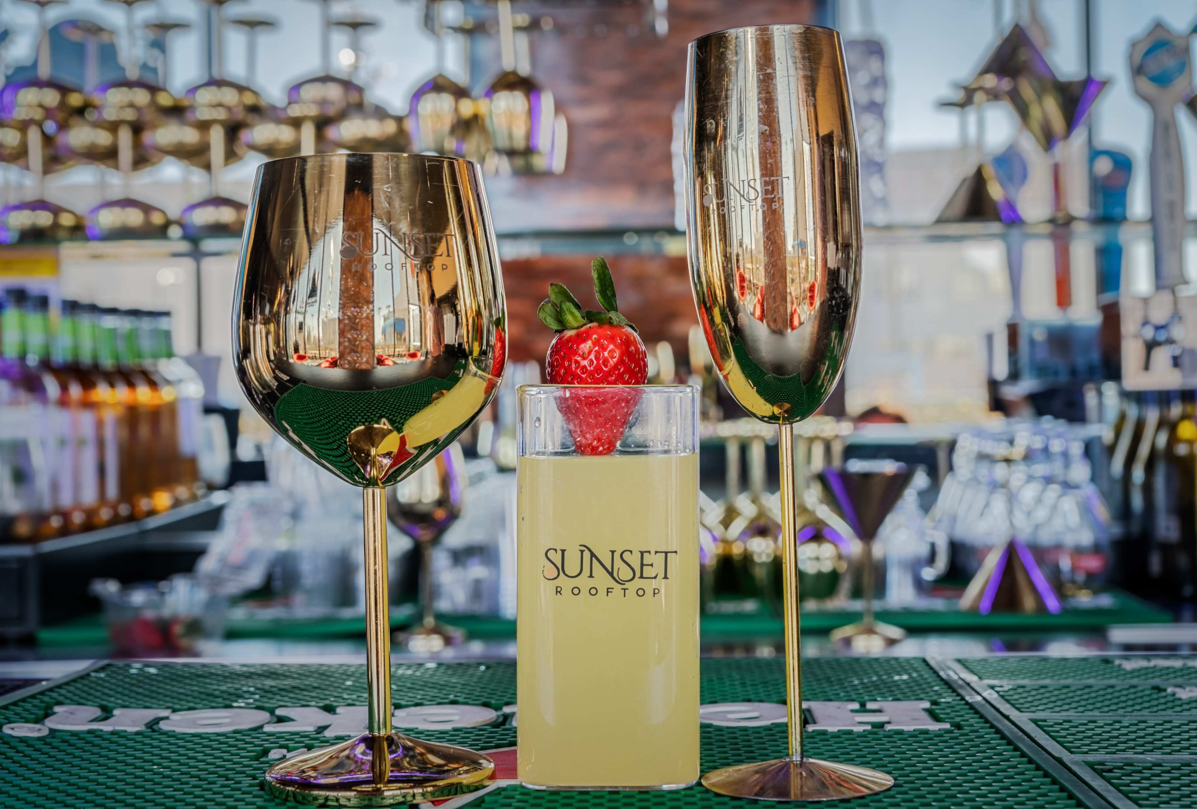 The image features two golden wine glasses flanking a short glass of yellow drink garnished with a strawberry, set on a green bar mat against a vibrant bar backdrop.