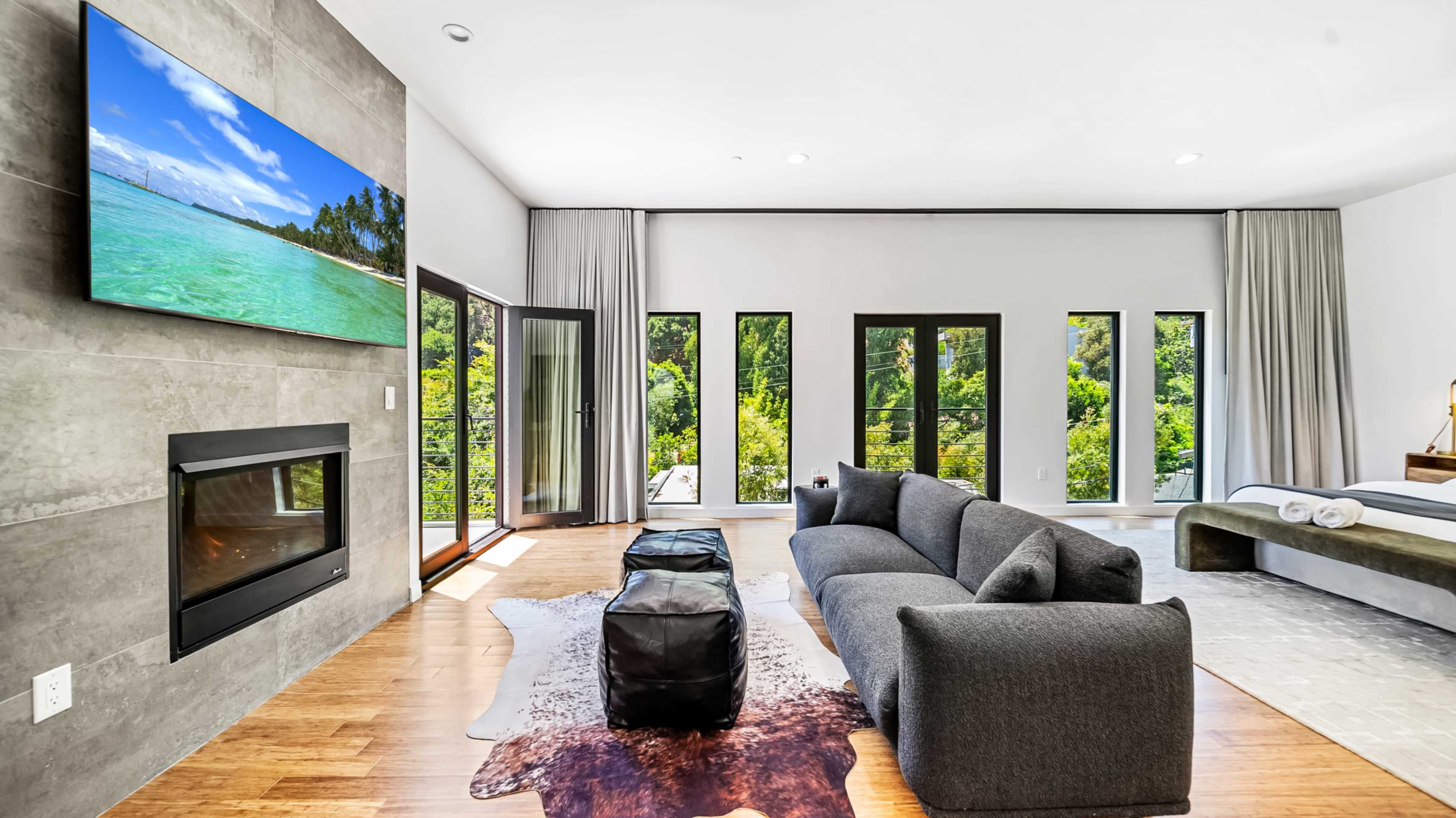 A modern living room features a large television mounted on a gray wall, a dark gray sofa, and floor-to-ceiling windows offering a view of greenery outside.