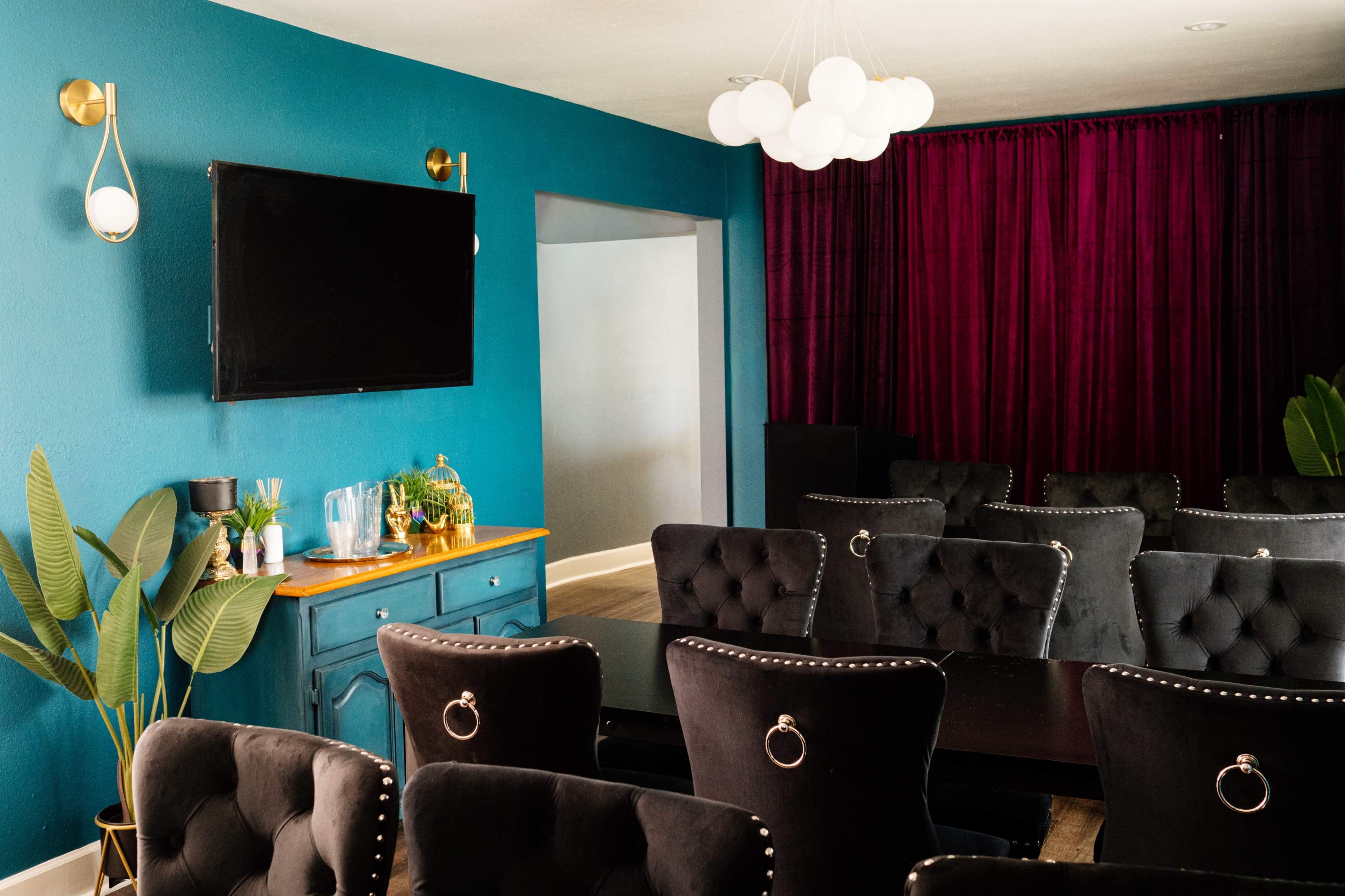 A modern conference room features a blue wall, a black table surrounded by tufted chairs, and a large television mounted above a small bar area.