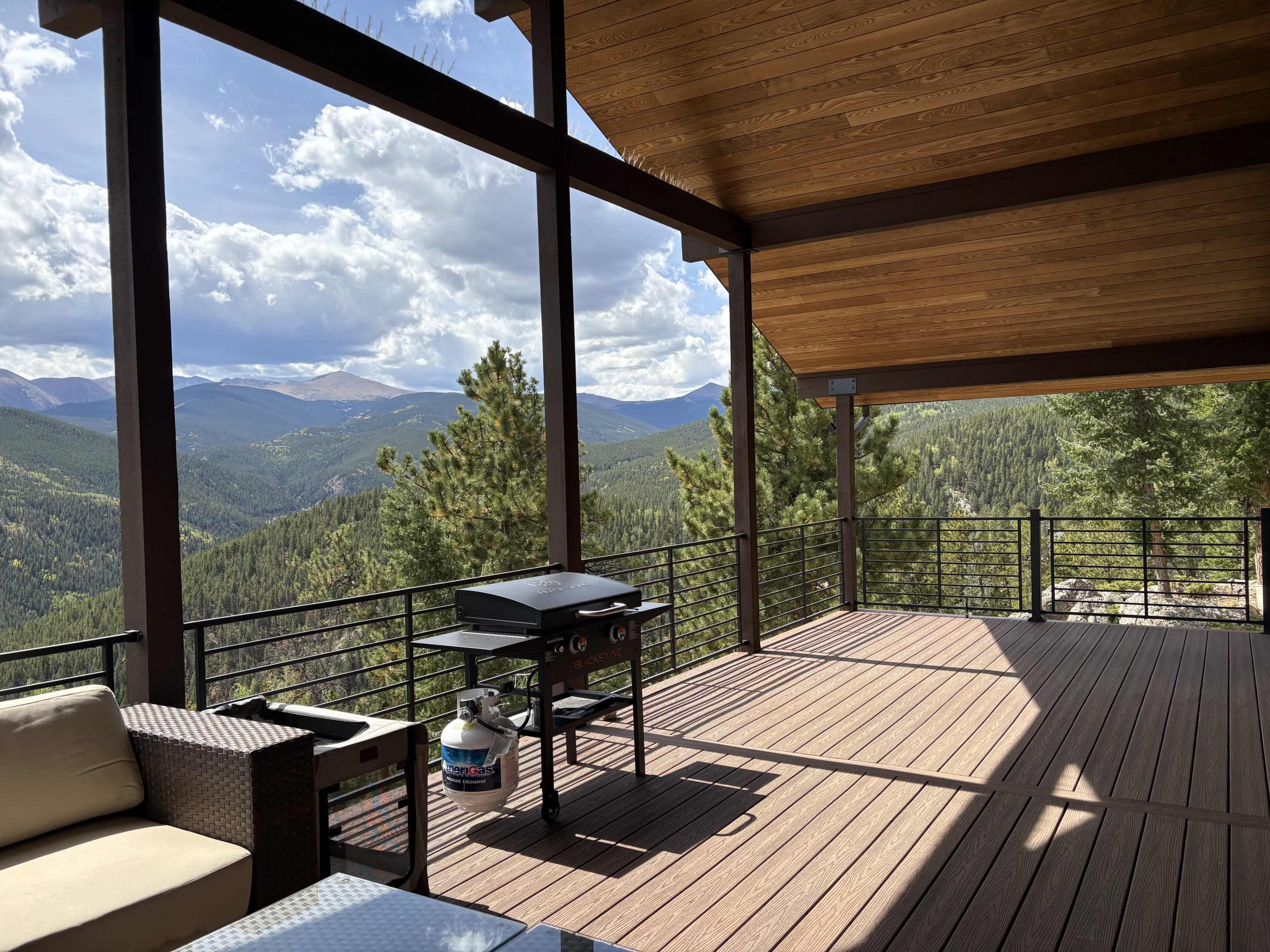 The image shows a spacious wooden deck with a grill overlooking a mountainous landscape under a partly cloudy sky.