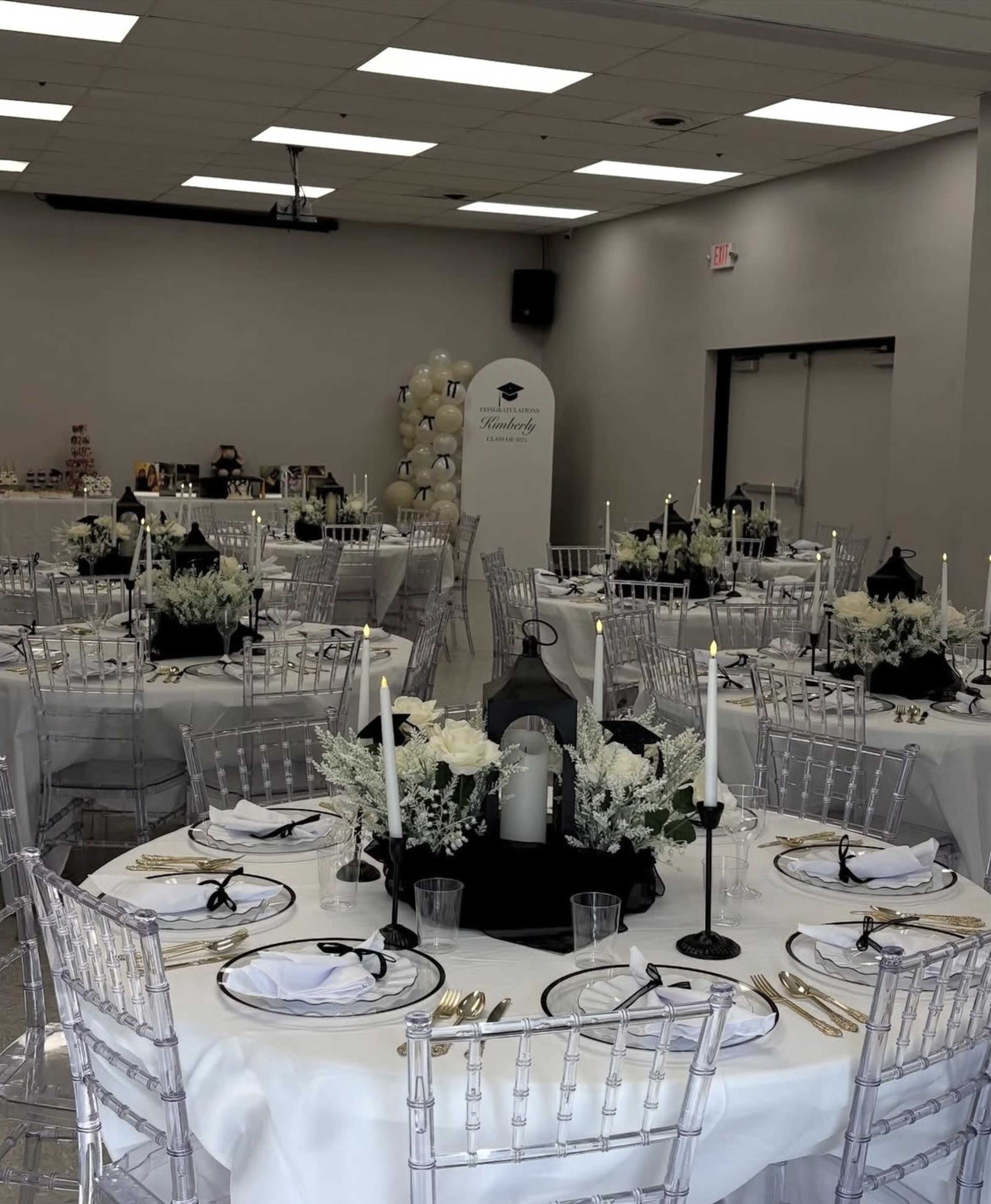 The image shows an elegantly arranged banquet hall with round tables set for a formal event, featuring white tablecloths, centerpieces with flowers, black accents, and decorative lanterns.