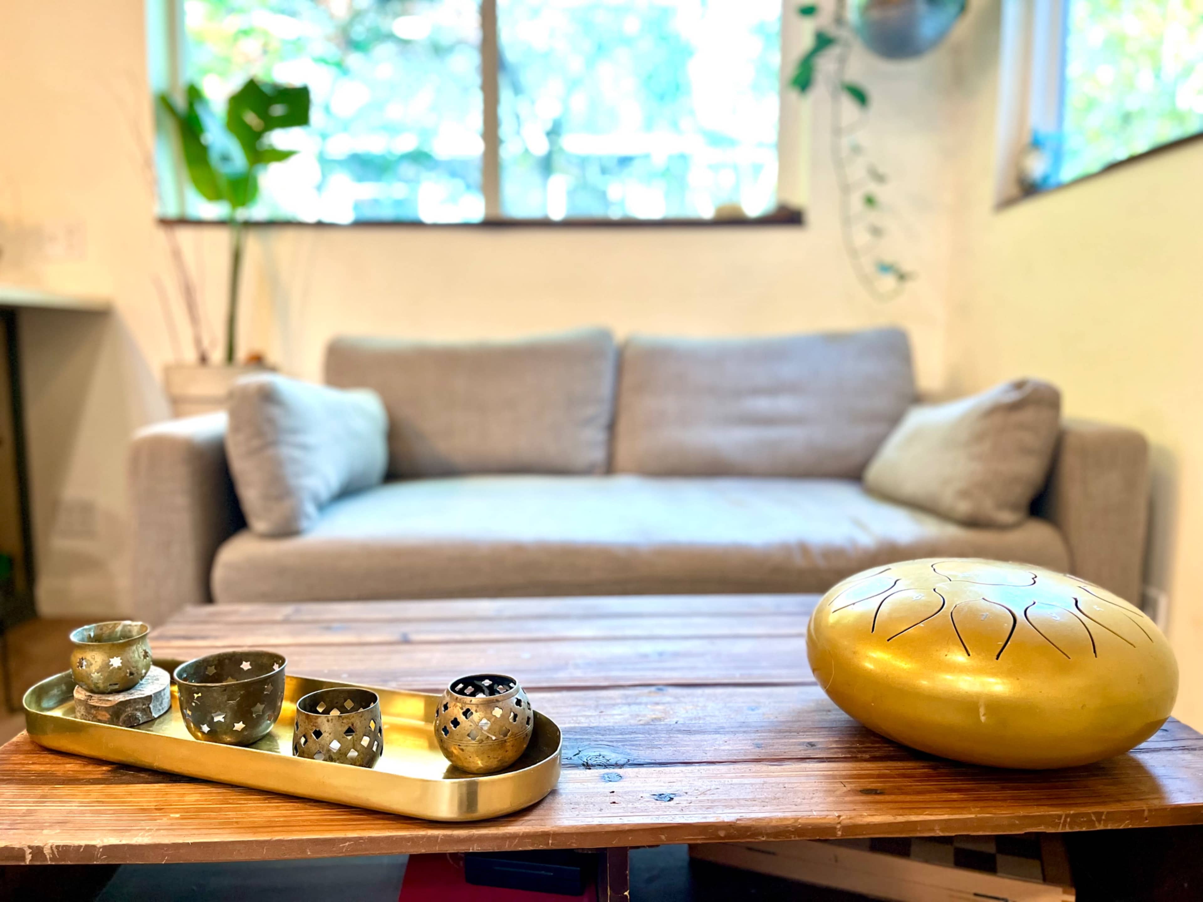 A wooden coffee table holds a tray with five decorative metal bowls, while a gray sofa is positioned in the background near a window with greenery.