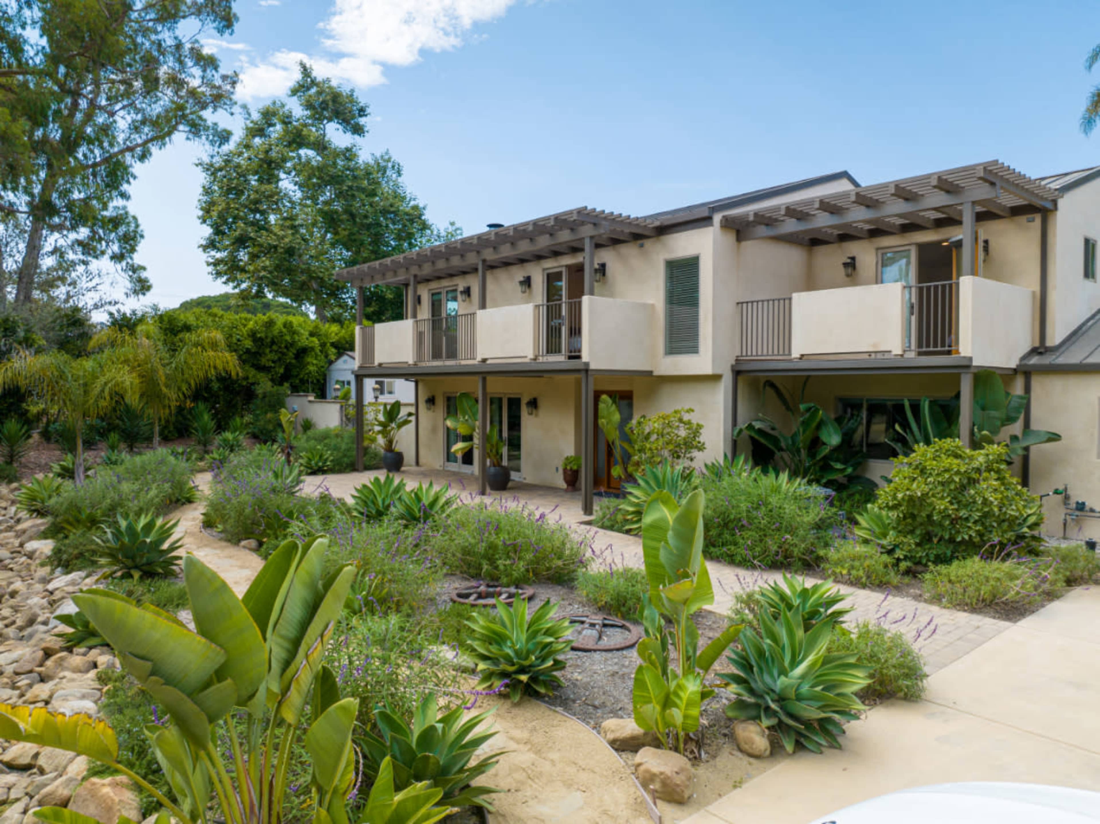 A modern two-story house with balconies overlooks a landscaped garden filled with various plants and rock formations.