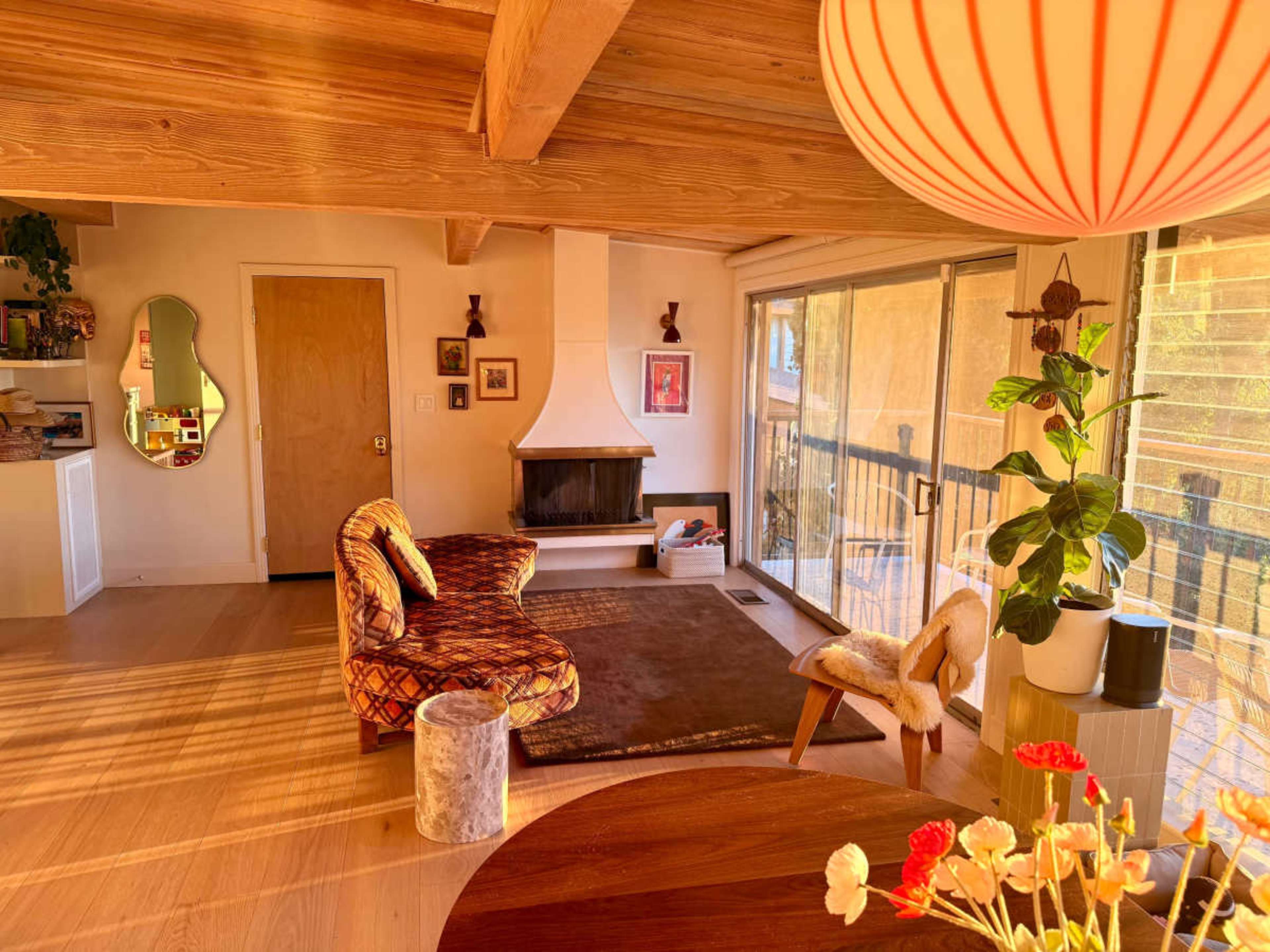 The image depicts a cozy living room with wooden beams, a fireplace, a large window, and a round dining table adorned with flowers.