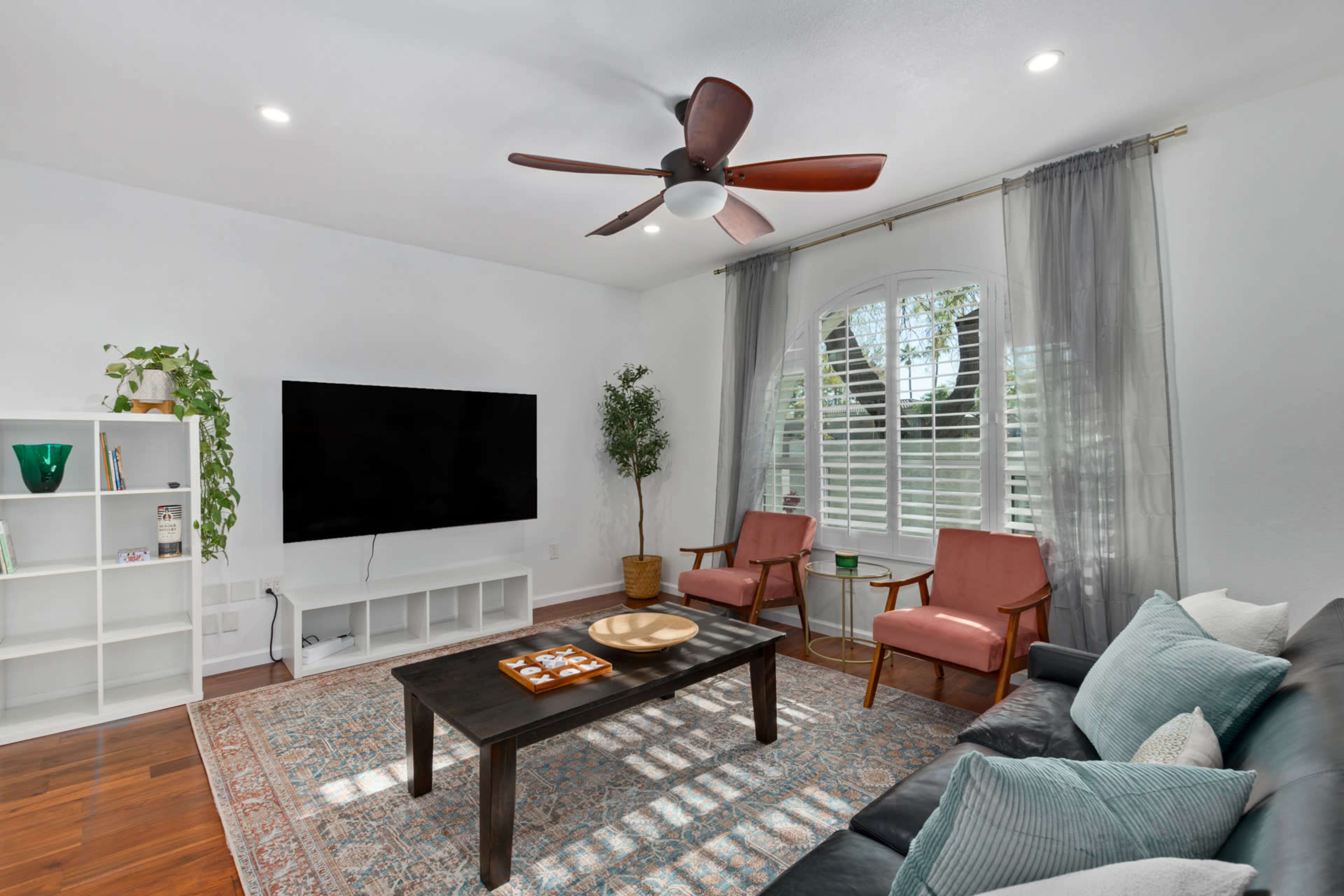 The image shows a modern living room with a flat-screen TV on the wall, two pink armchairs, a dark wooden coffee table, and a light-colored sofa, all arranged around a patterned area rug.
