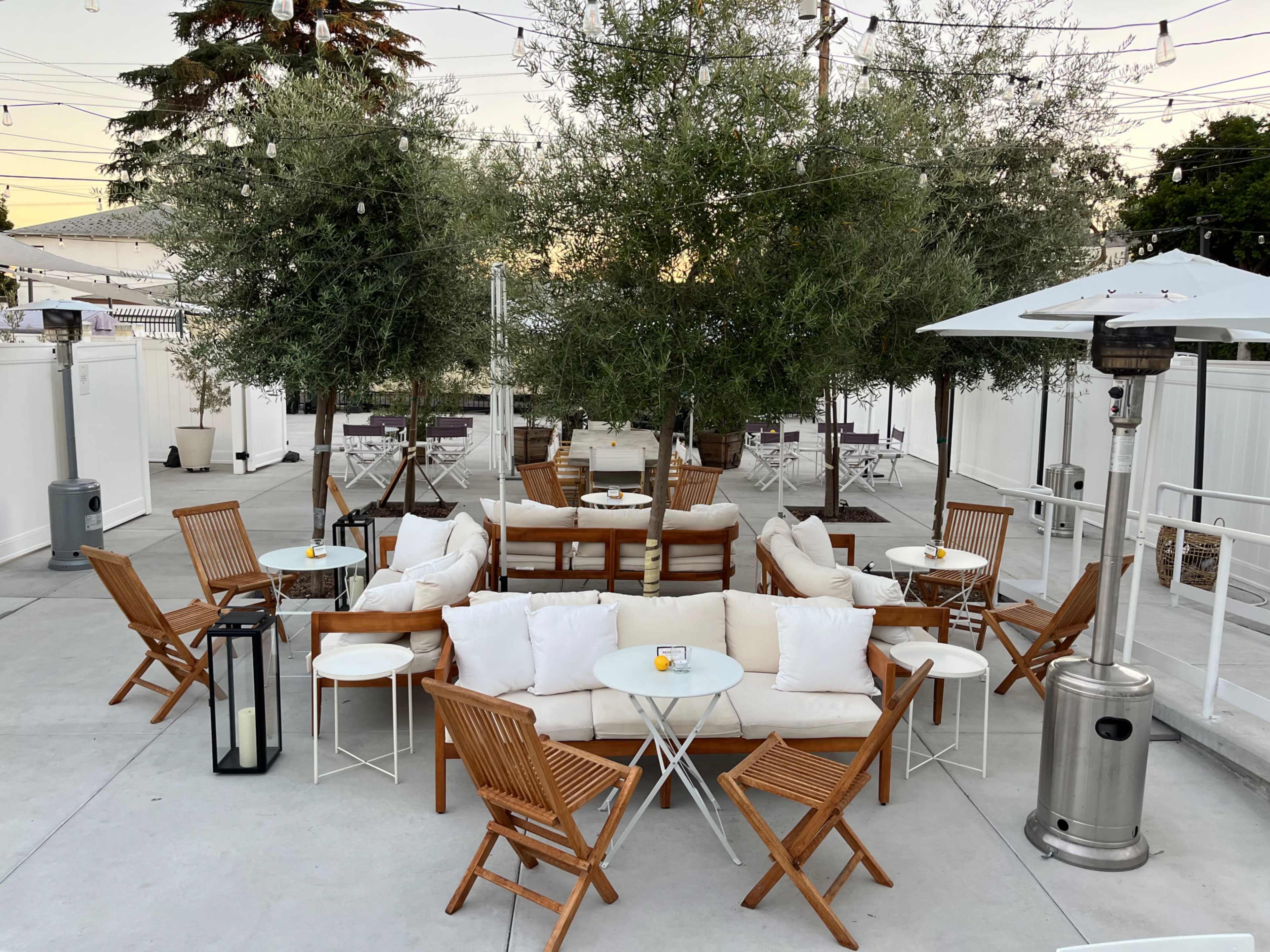A landscaped outdoor seating area features wooden chairs and tables arranged around olive trees, with patio lights overhead and gas heaters positioned along the perimeter.