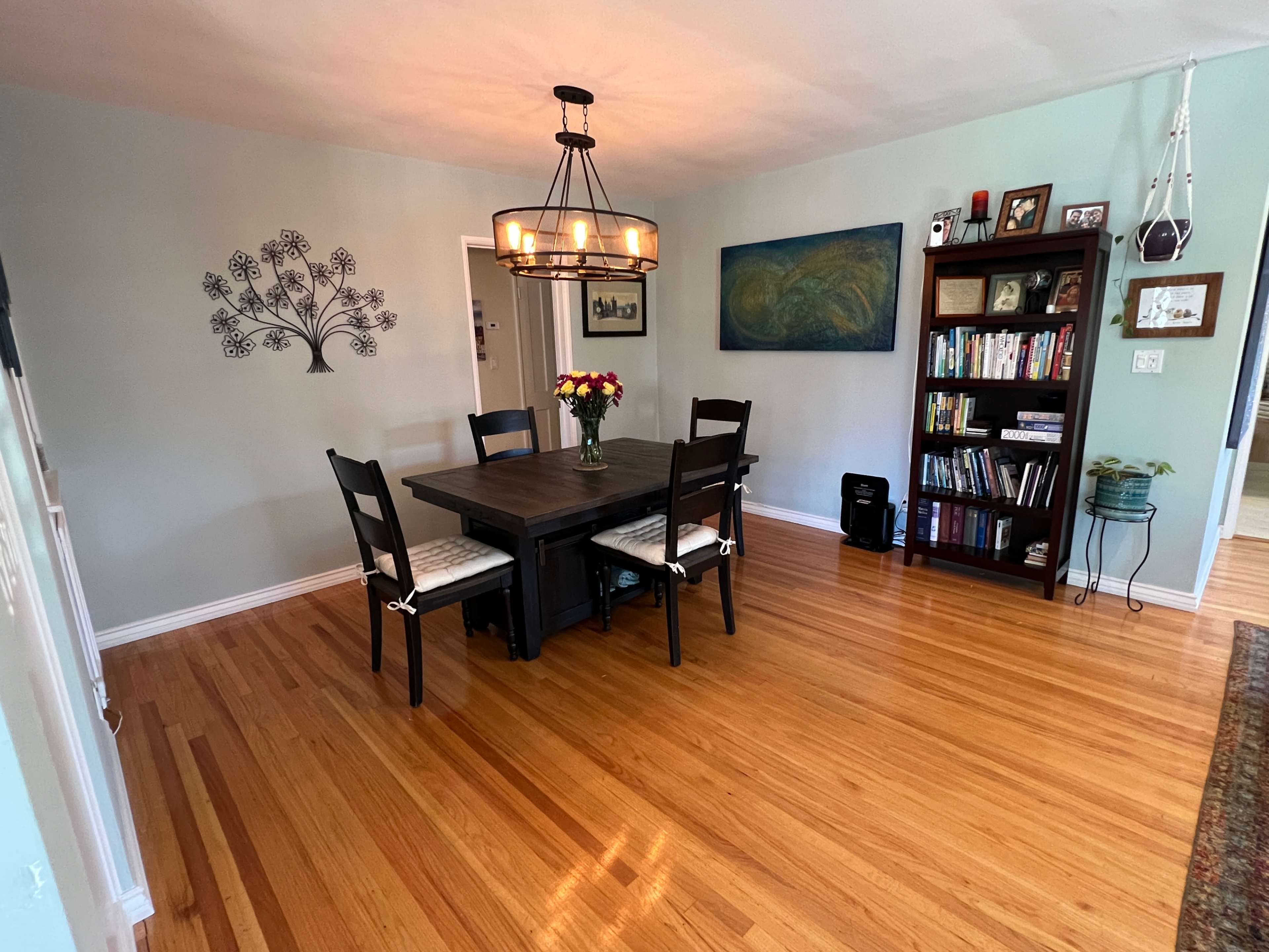 A dining area features a wooden table surrounded by chairs, with a chandelier above, a bookshelf filled with books, and a decorative wall design.