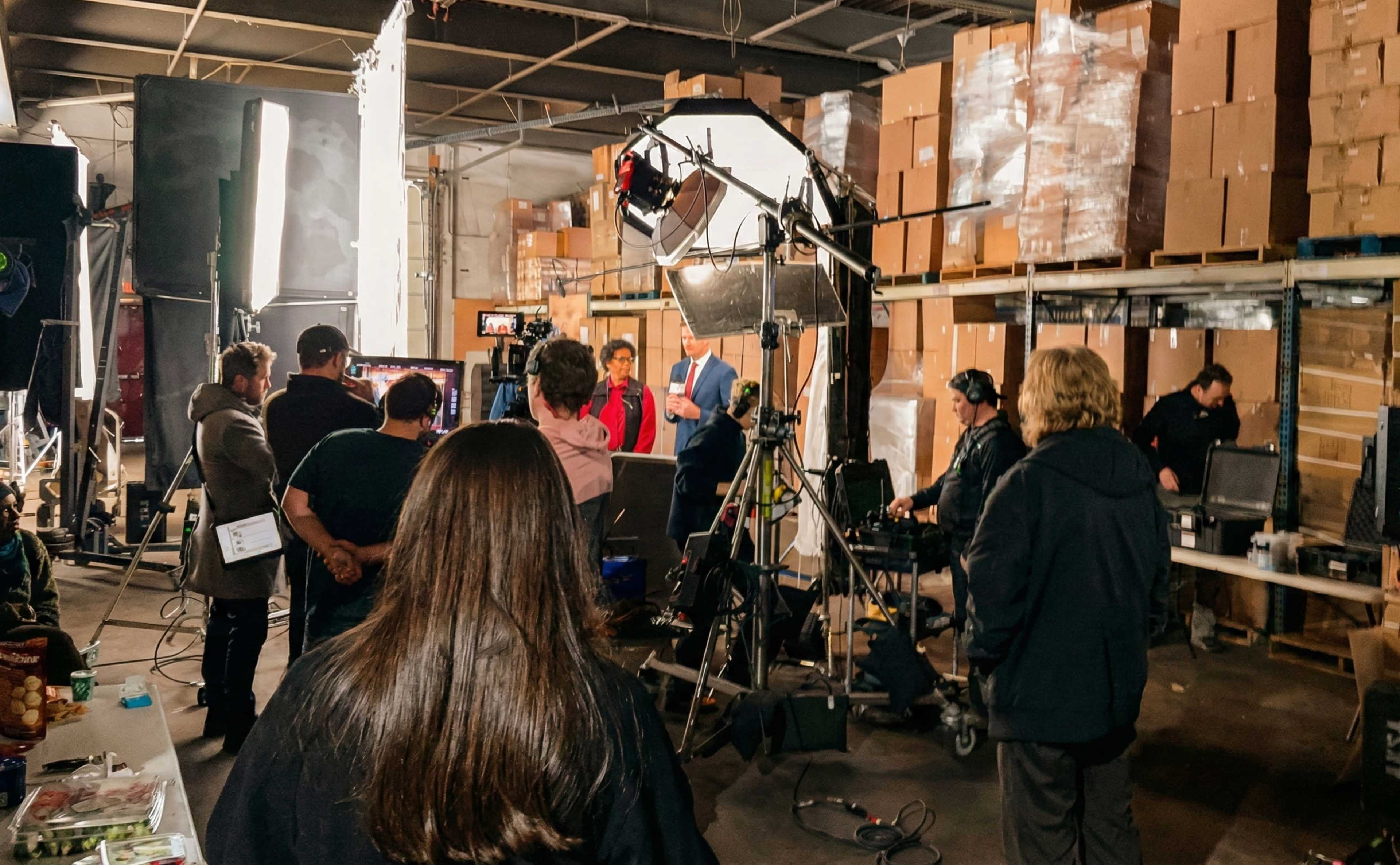 A film crew is set up in a warehouse filled with stacked boxes, working around lights and cameras while actors prepare for a scene.
