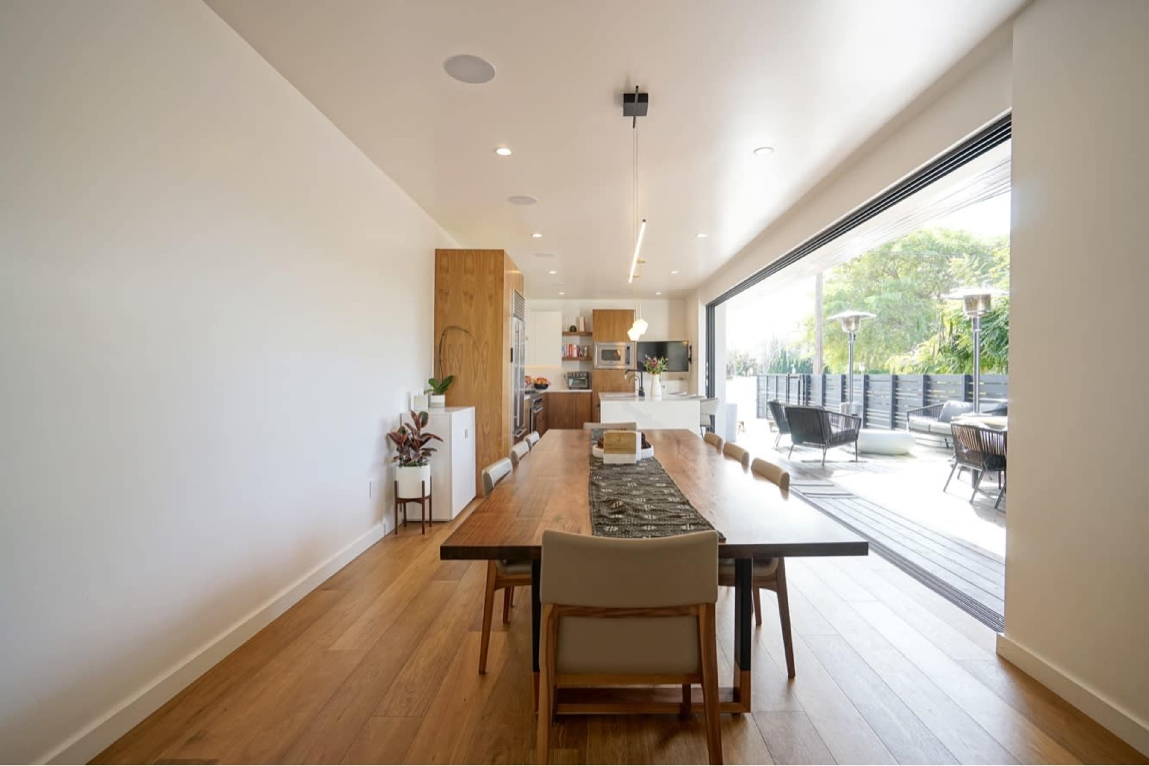 The image shows a modern dining area with a long wooden table, surrounded by chairs, leading to an open kitchen and outdoor patio through large glass doors.