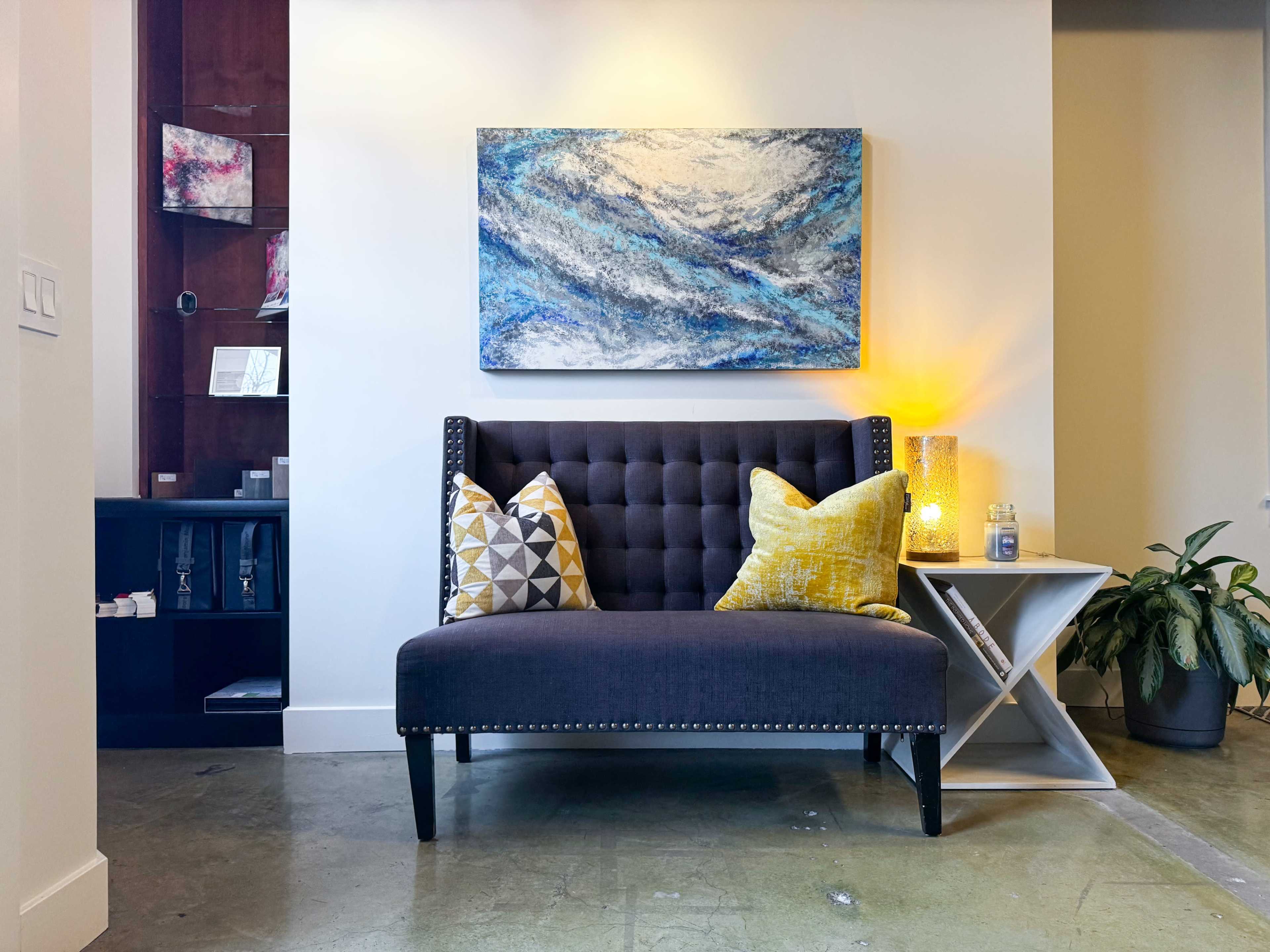 A tufted gray settee with decorative pillows is positioned against a light wall, accompanied by a geometric side table and a small plant in a corner.