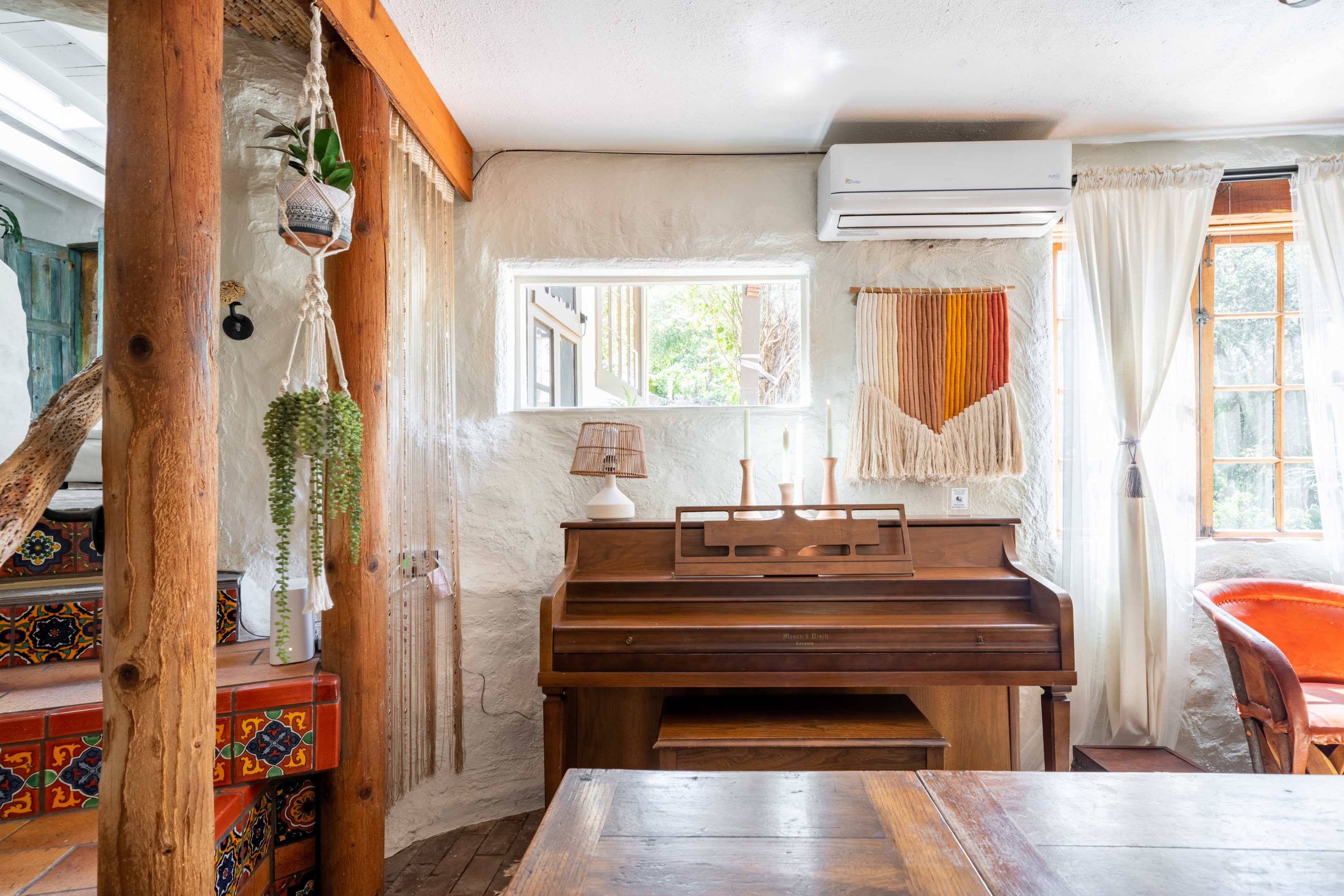 A wooden piano sits in a cozy room with natural light streaming through the windows, surrounded by decorative elements and wooden beams.