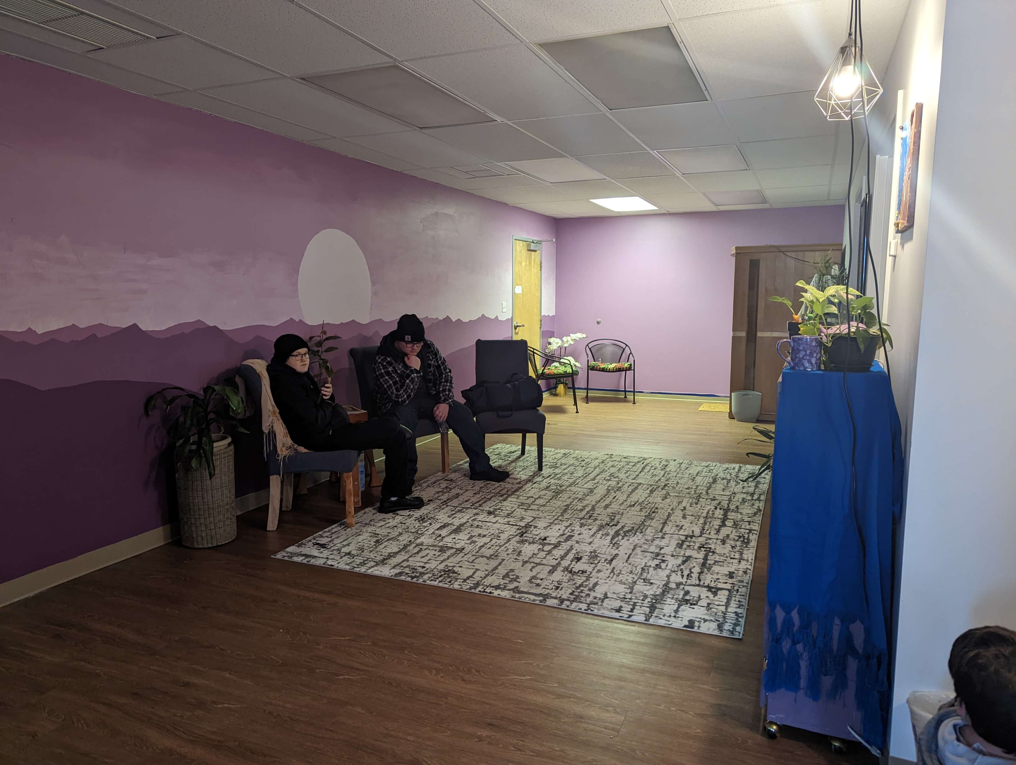 A purple-walled waiting area with two people sitting on chairs and a decorative rug on the floor.
