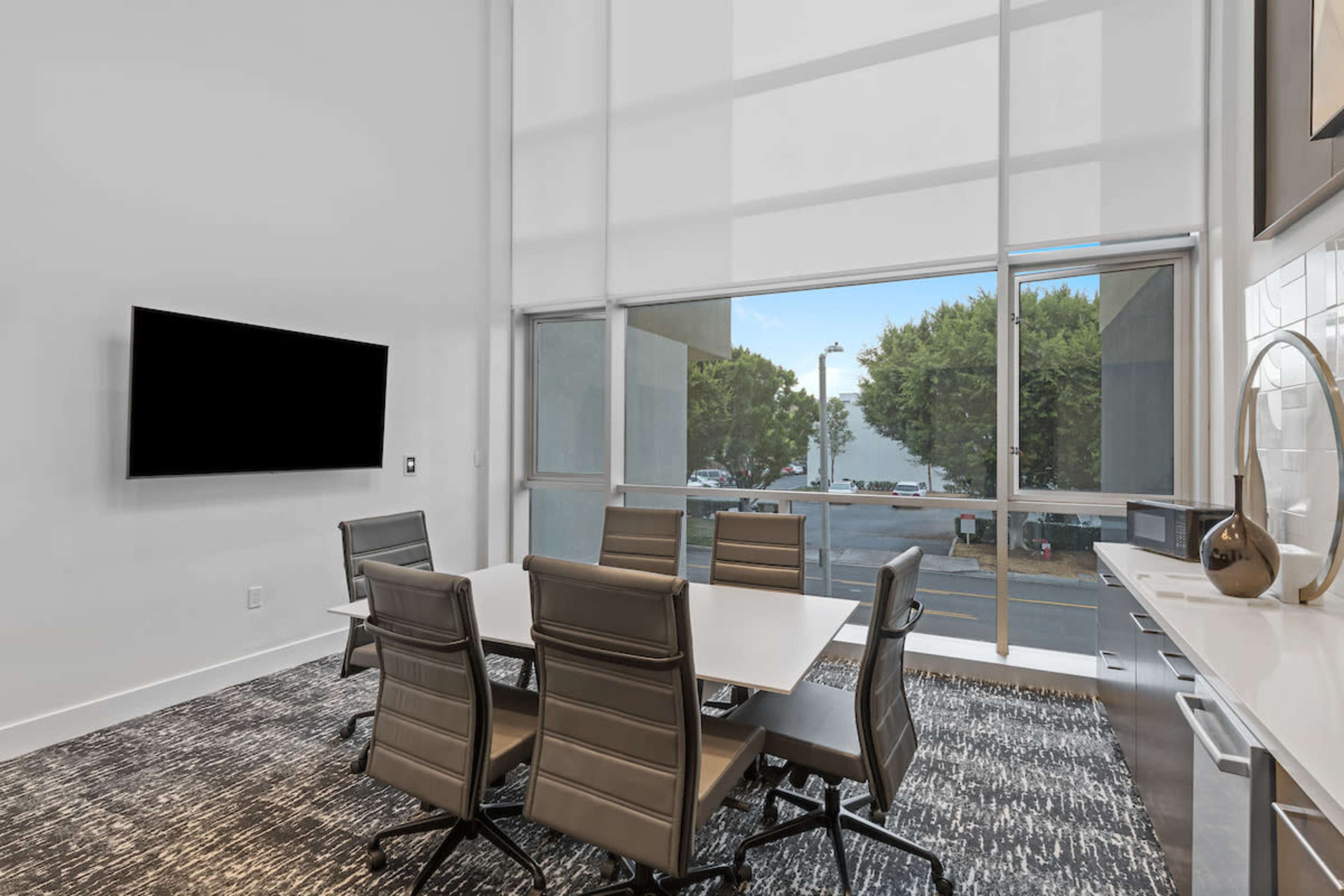 A modern conference room features a large window, a rectangular table surrounded by six leather chairs, and a wall-mounted television.