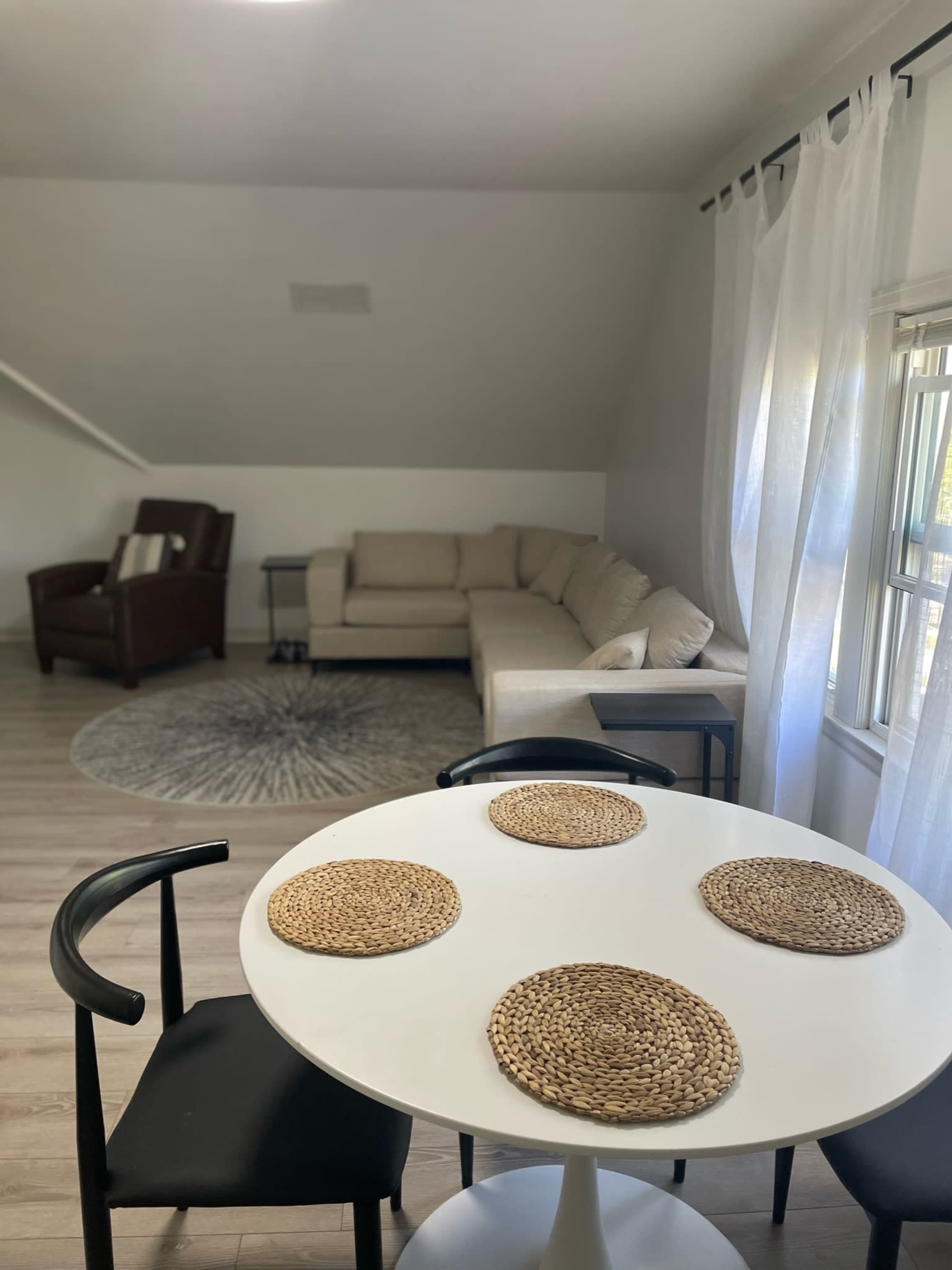 The image shows a well-lit living space featuring a dining table with four placemats, a circular rug, a sectional couch, and an armchair.