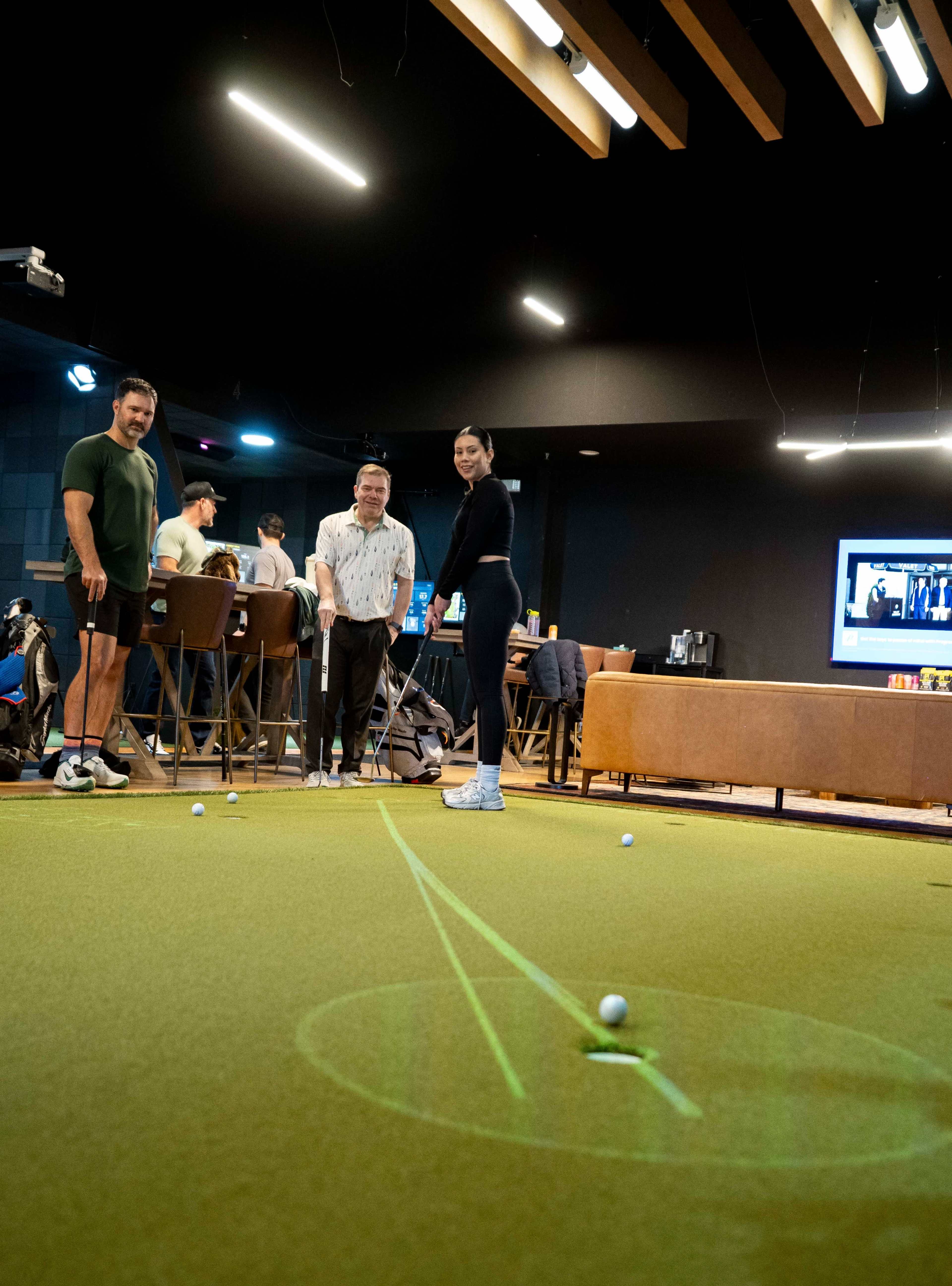 Three people are gathered around a putting green inside a venue, with one person preparing to take a shot while the others watch.