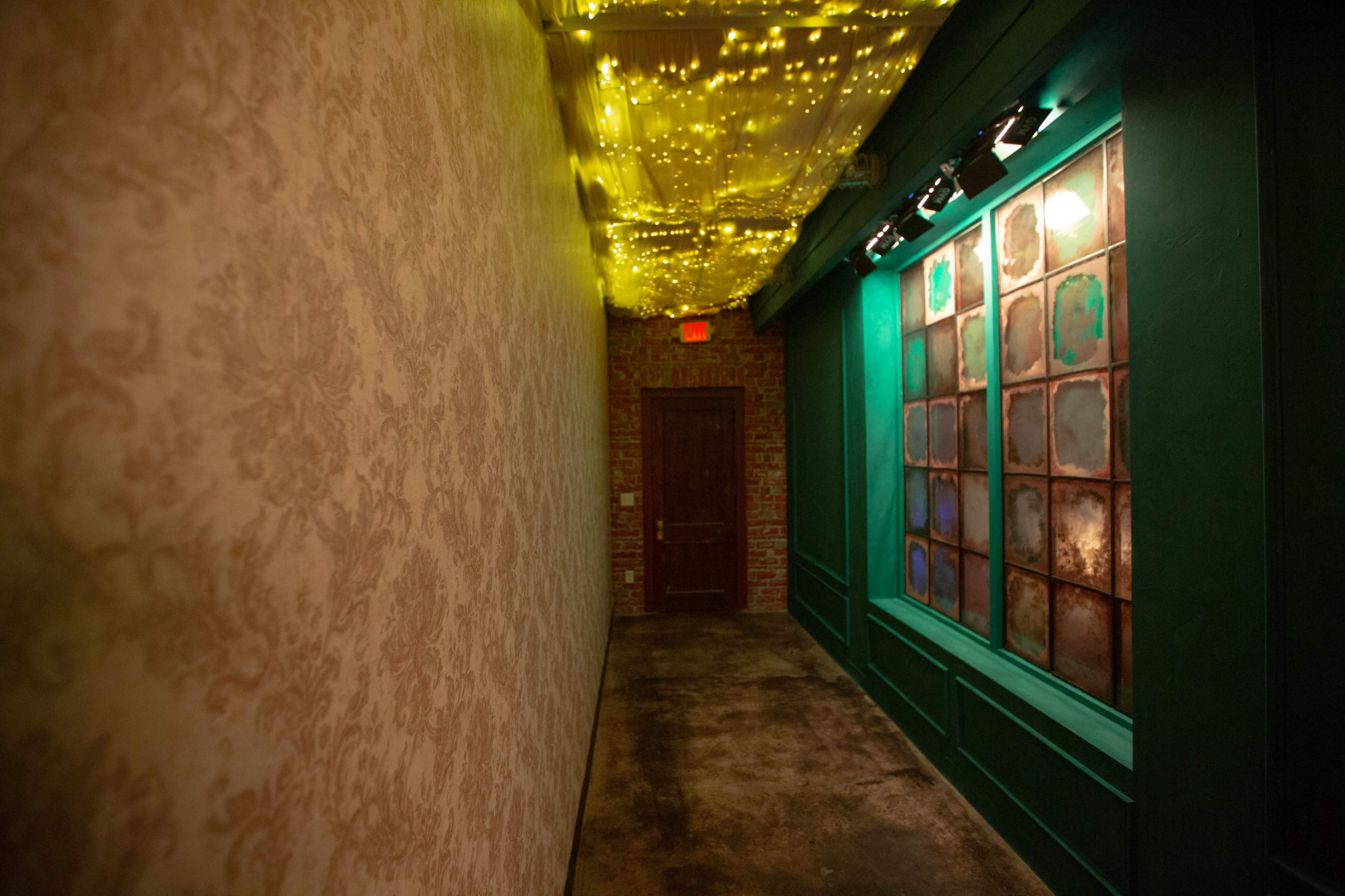 A narrow hallway features patterned wallpaper, a green wall with a decorative window, and string lights illuminating the ceiling.