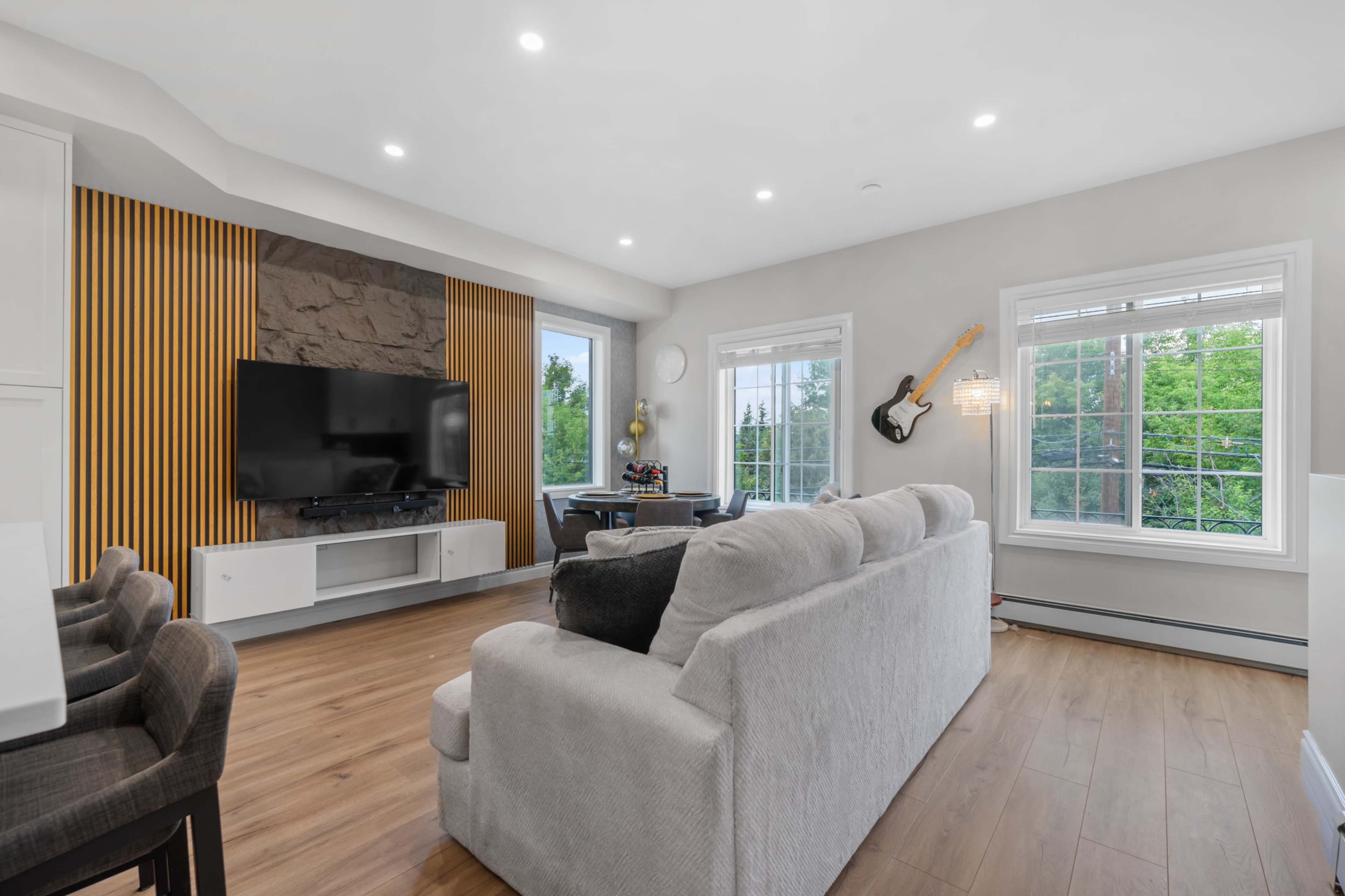 The living room features a television mounted on a stone wall, with large windows overlooking greenery and a guitar hanging on the wall.